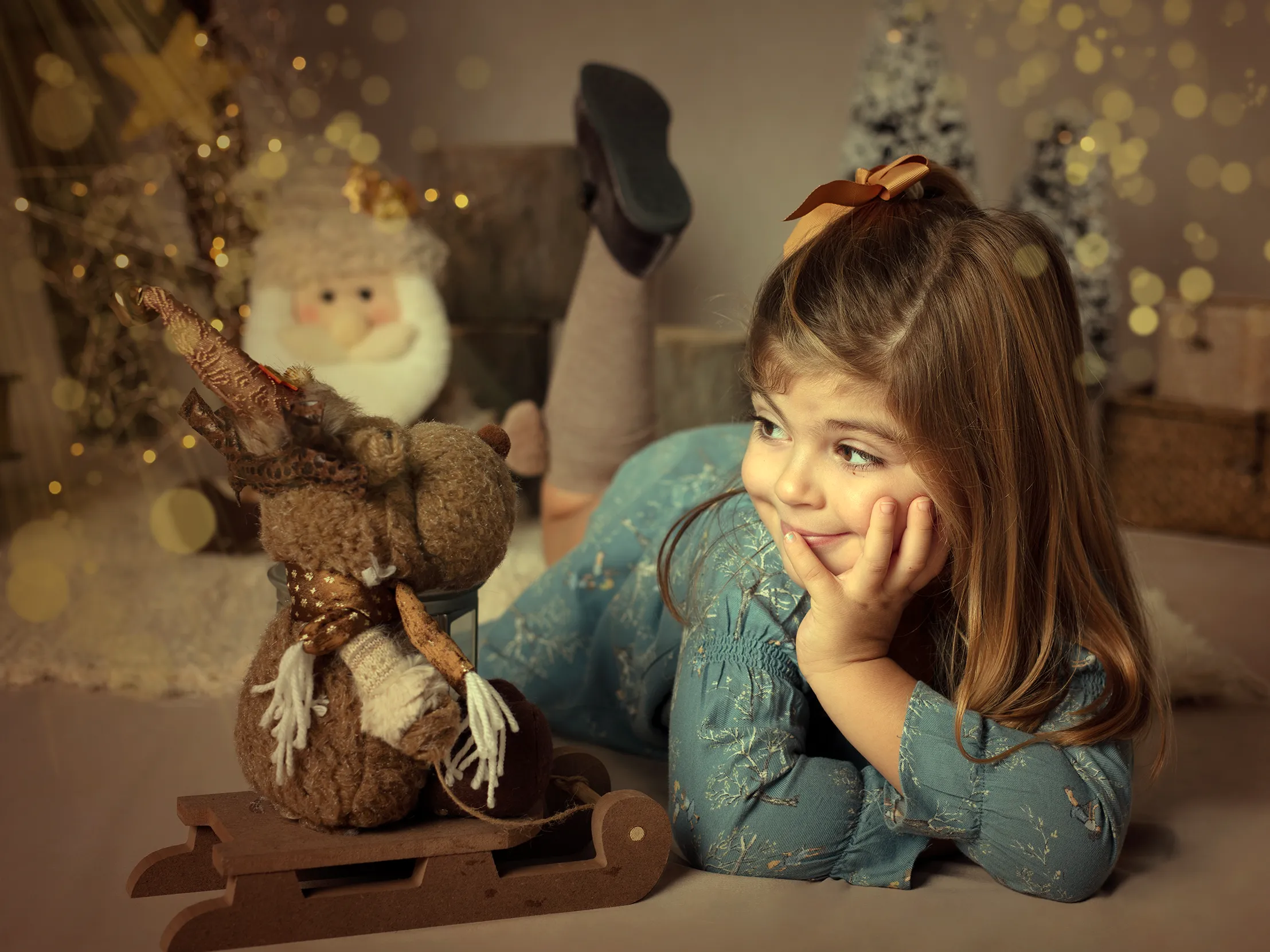 Una niña pequeña con vestido azul sonríe, apoyándose en la mano con un reno de juguete y adornos navideños.
