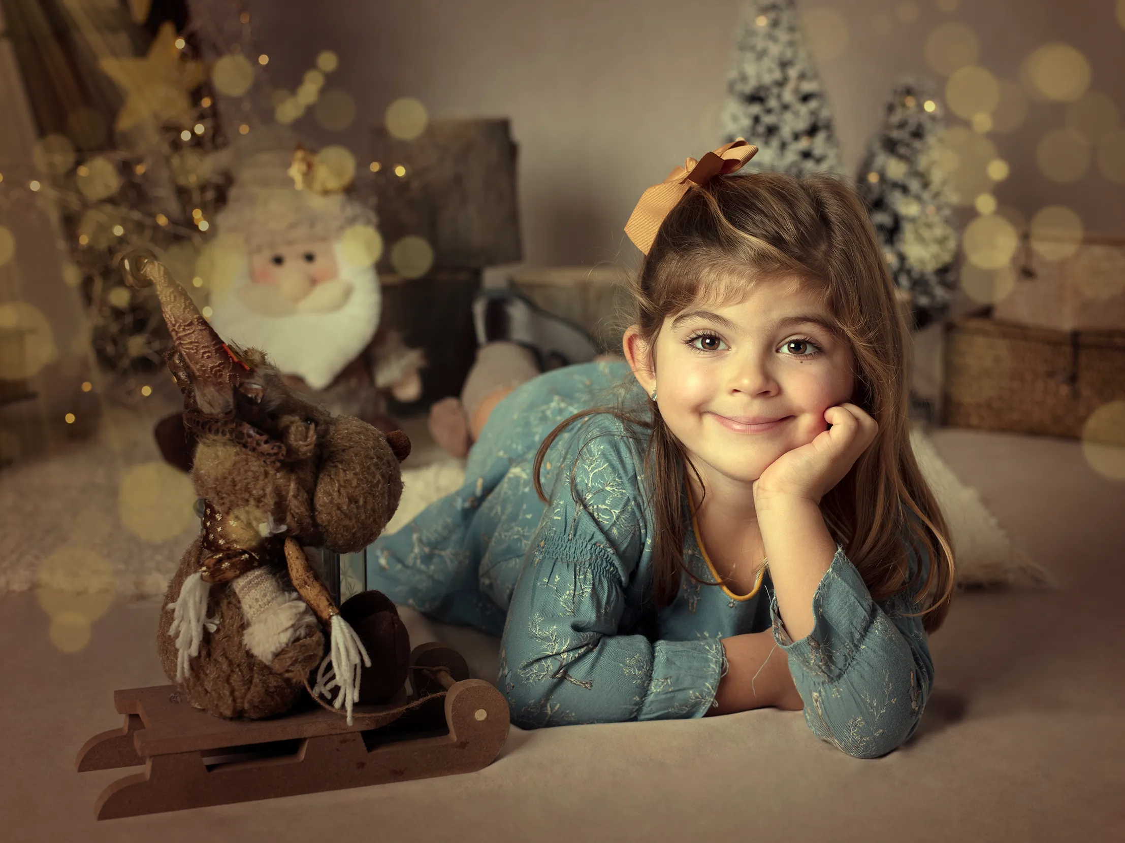 Una niña pequeña sonríe, recostada en una escena navideña con renos y Papá Noel.