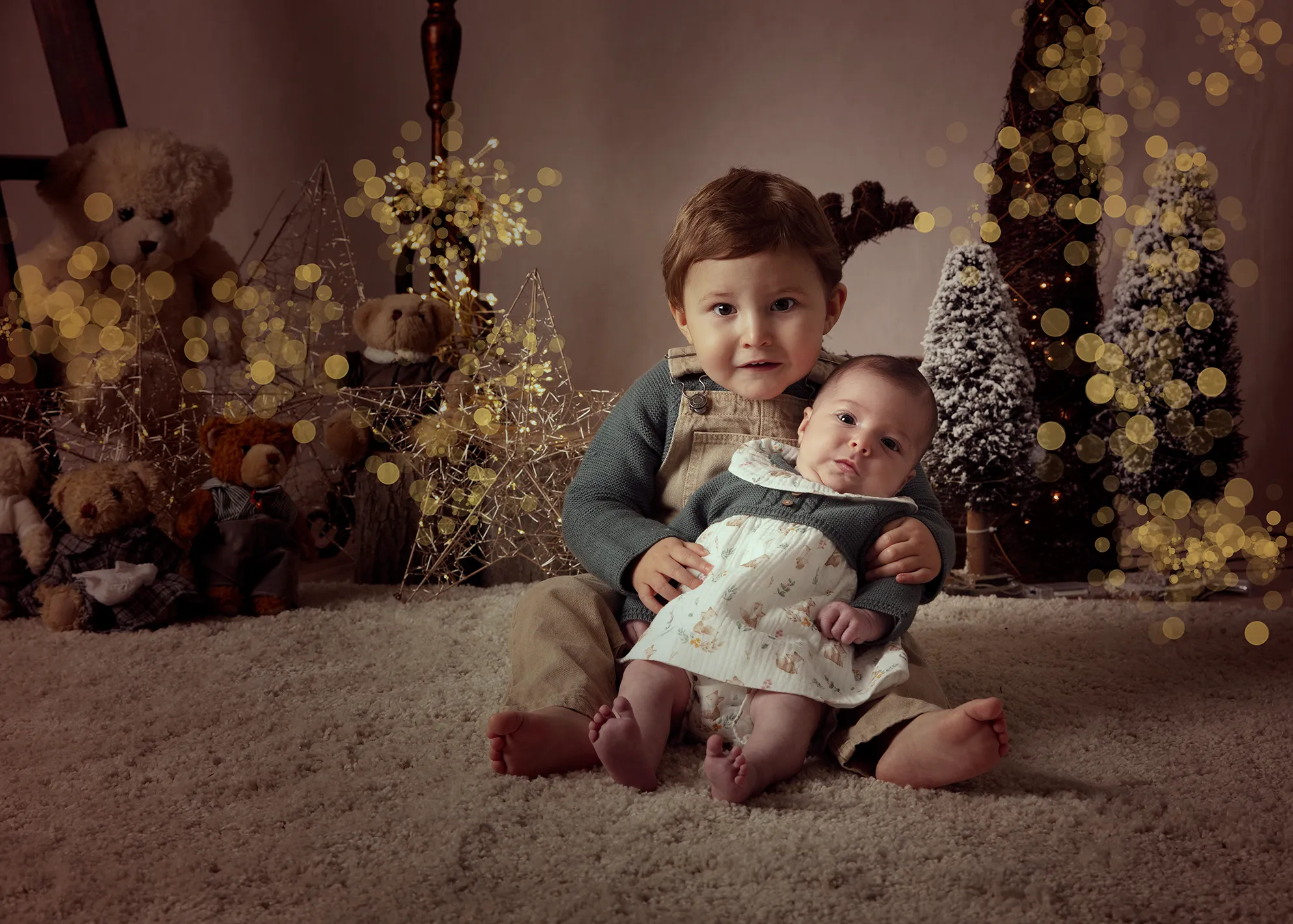 Niño con cuernos de reno sosteniendo a un bebé; ambos sonriendo, en un ambiente navideño con ositos de peluche y luces.