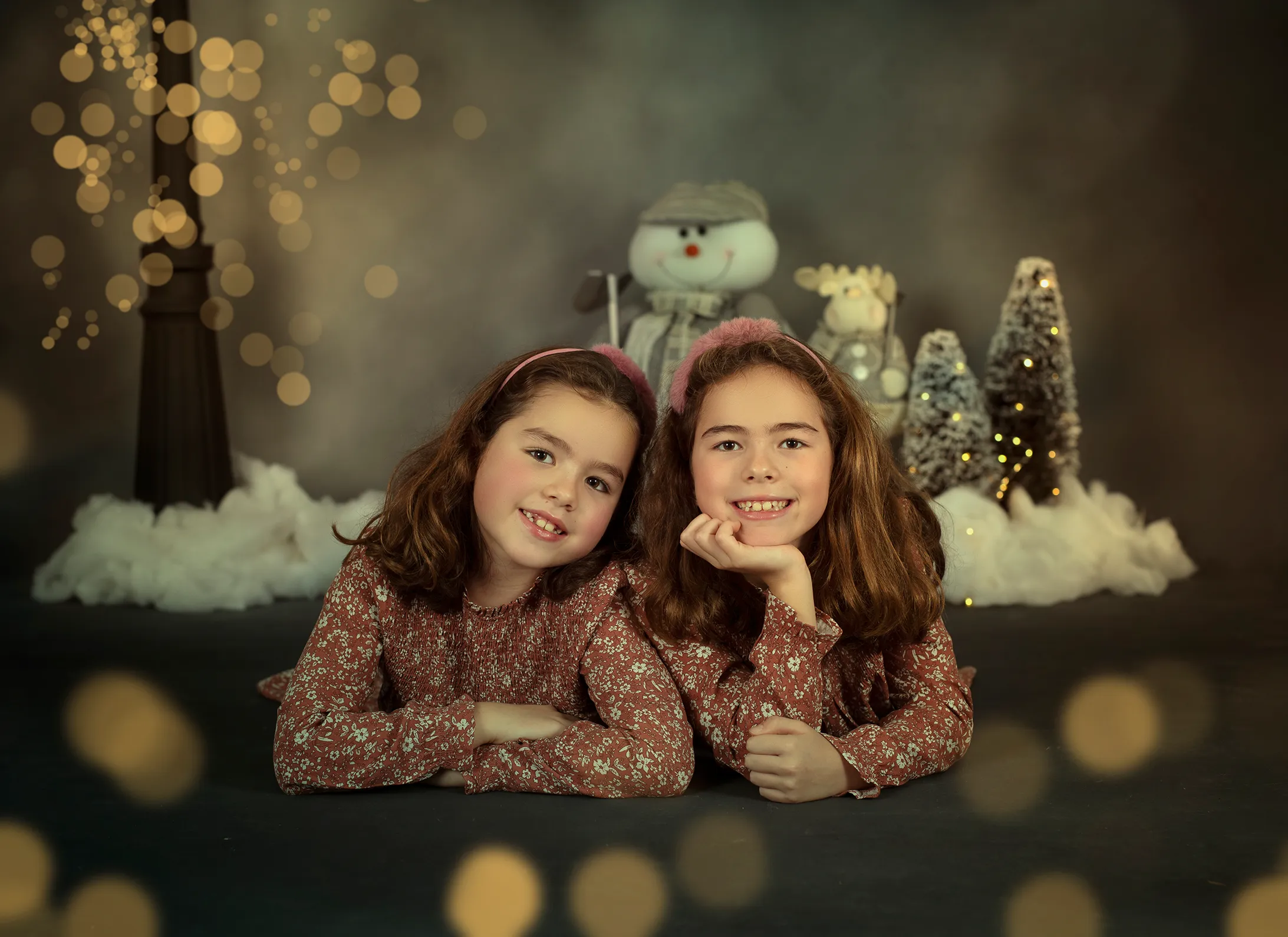 Dos gemelas posan sonrientes en un paisaje invernal. Al fondo, un muñeco de nieve y árboles de Navidad.
