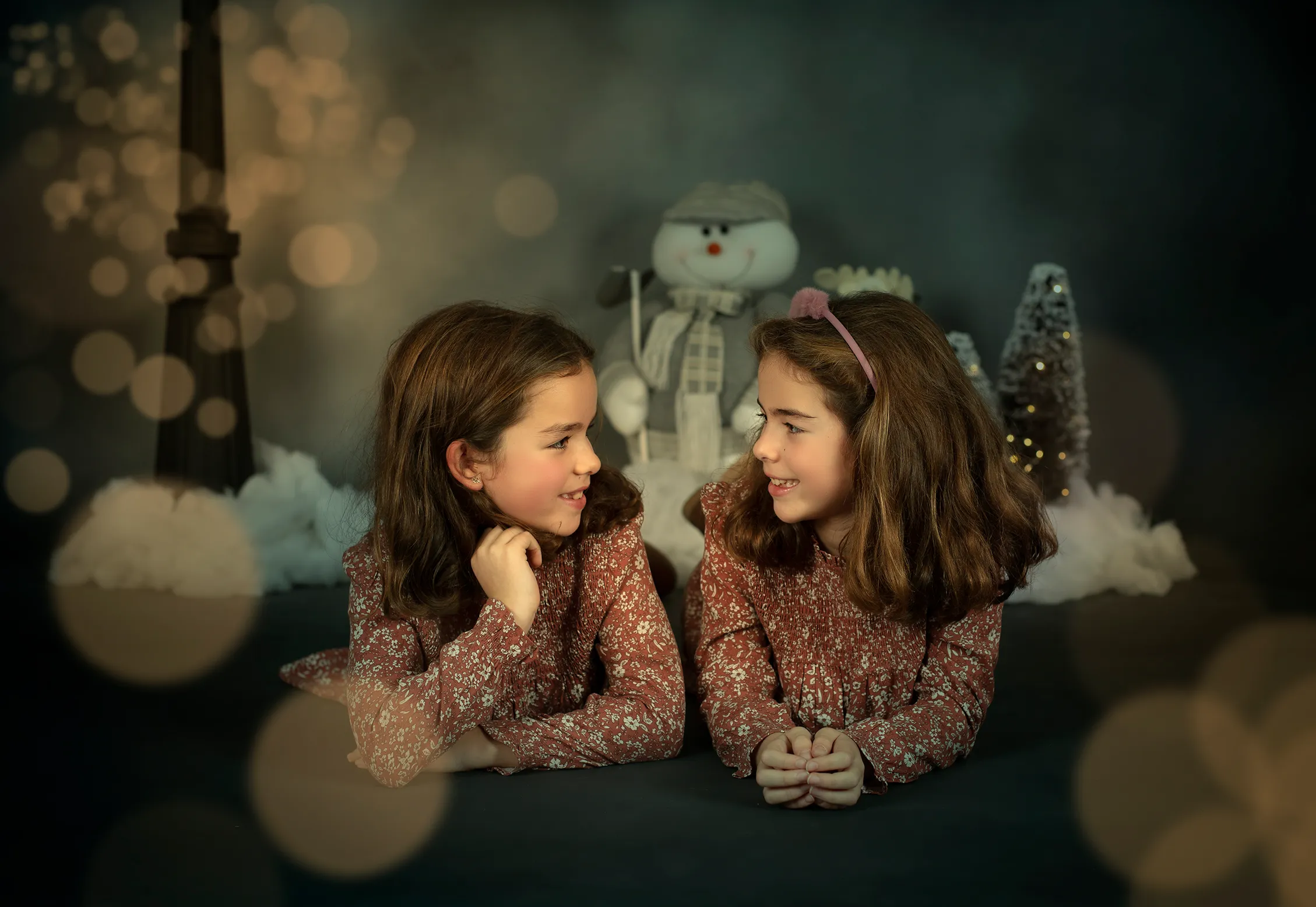 Dos chicas con suéteres marrones se miran fijamente en una escena invernal con un muñeco de nieve y efecto bokeh.