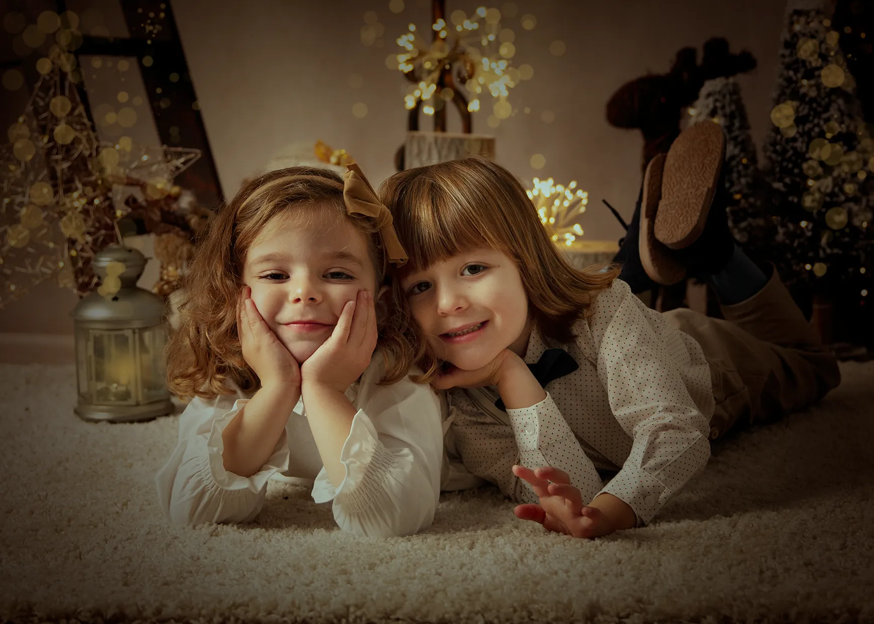 Dos niños, sonrientes, tumbados sobre una alfombra. Escenario navideño decorado con luces de fondo.