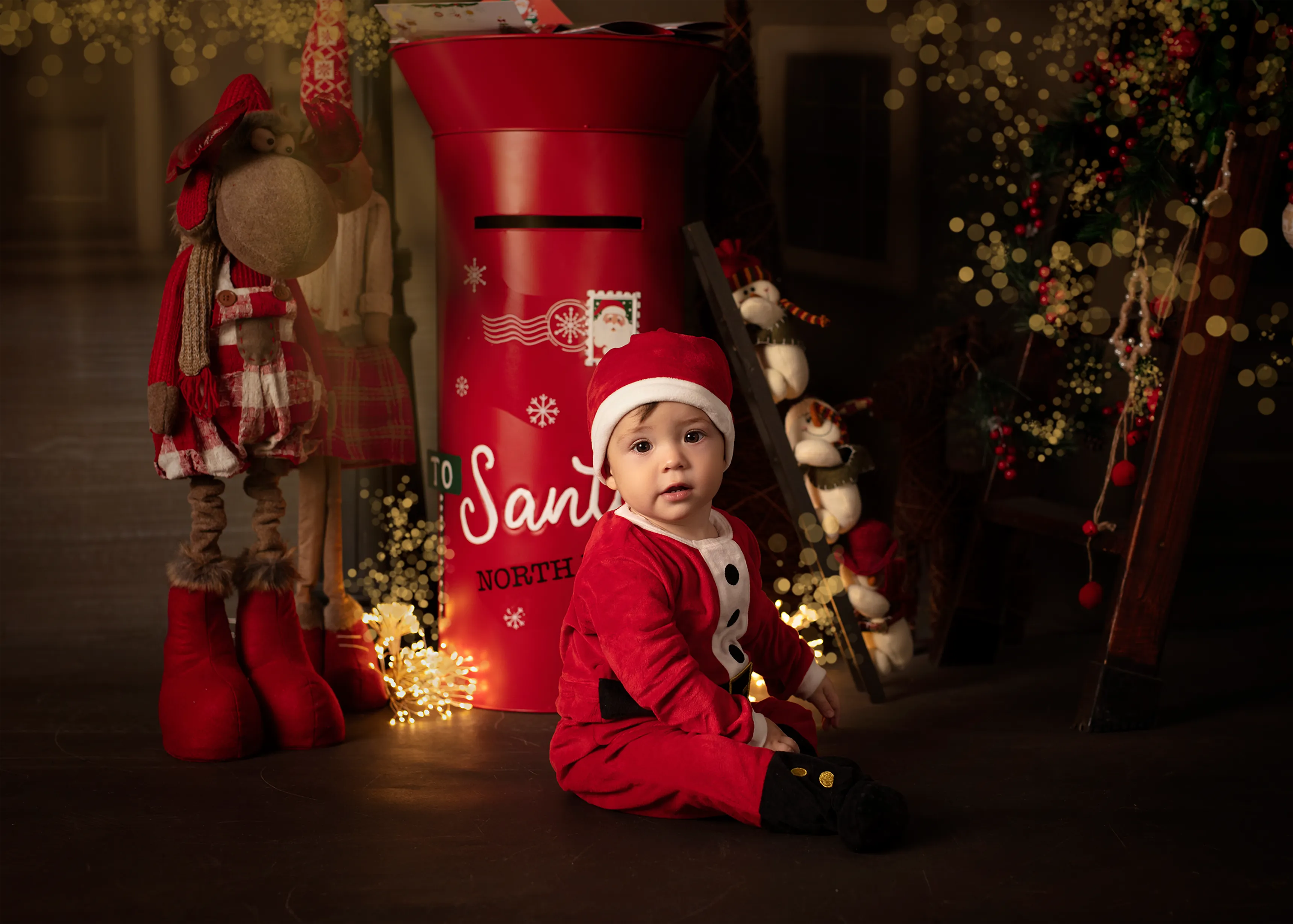 Un niño vestido de Papá Noel está sentado cerca de un buzón rojo y adornos navideños.