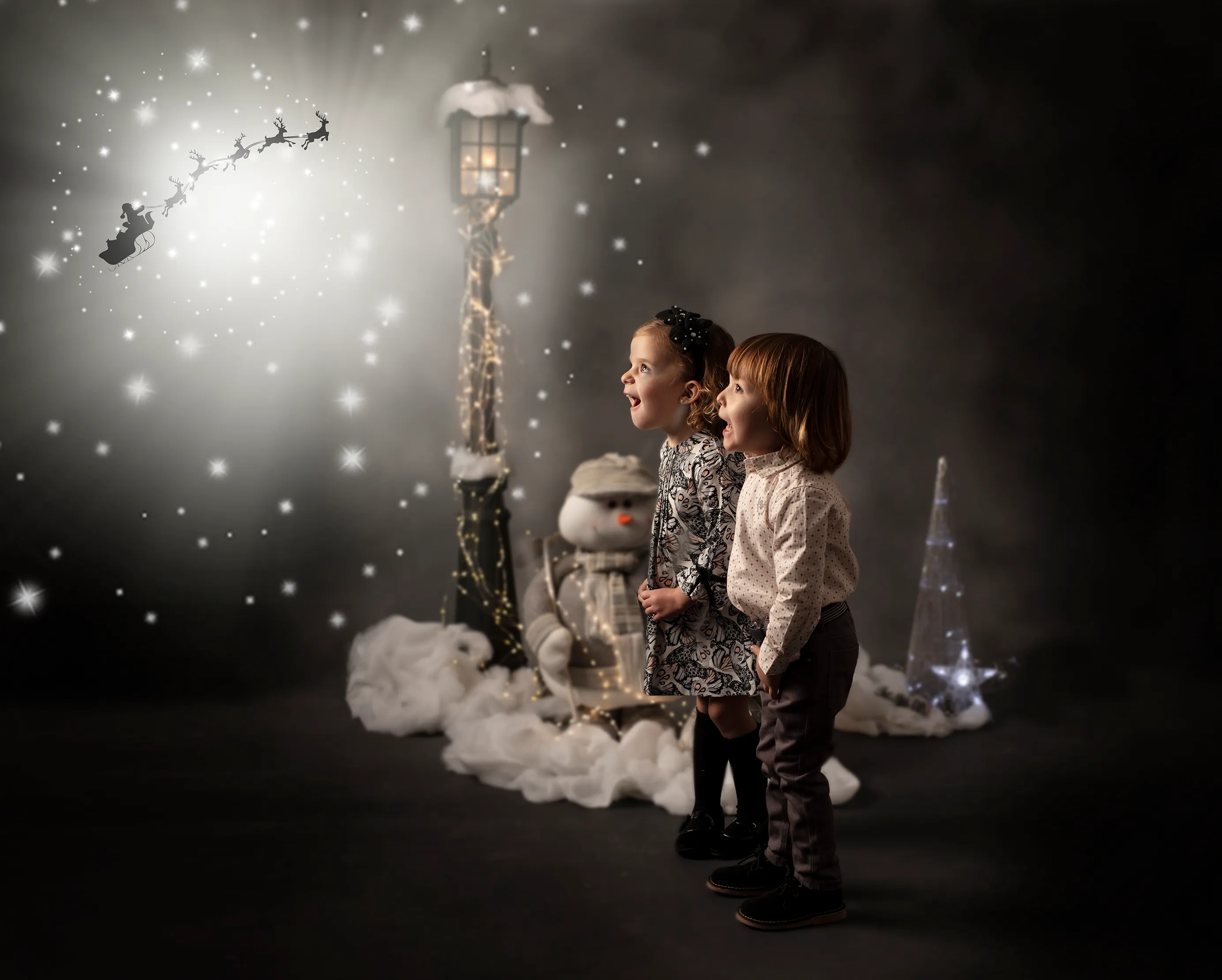 Dos niños miran hacia arriba, hacia el trineo de Papá Noel, mientras caen copos de nieve. Farola festiva y escena nevada.