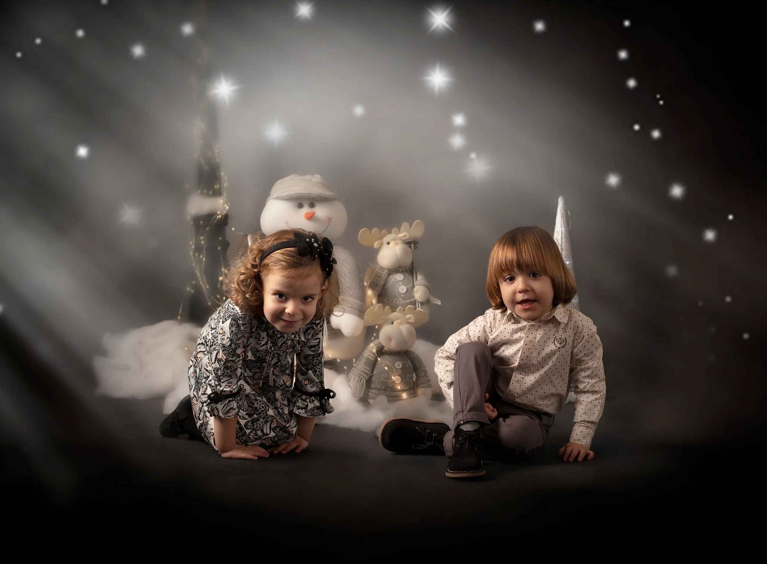 Dos niños están sentados frente a un muñeco de nieve y decoraciones, con efecto de nieve cayendo.