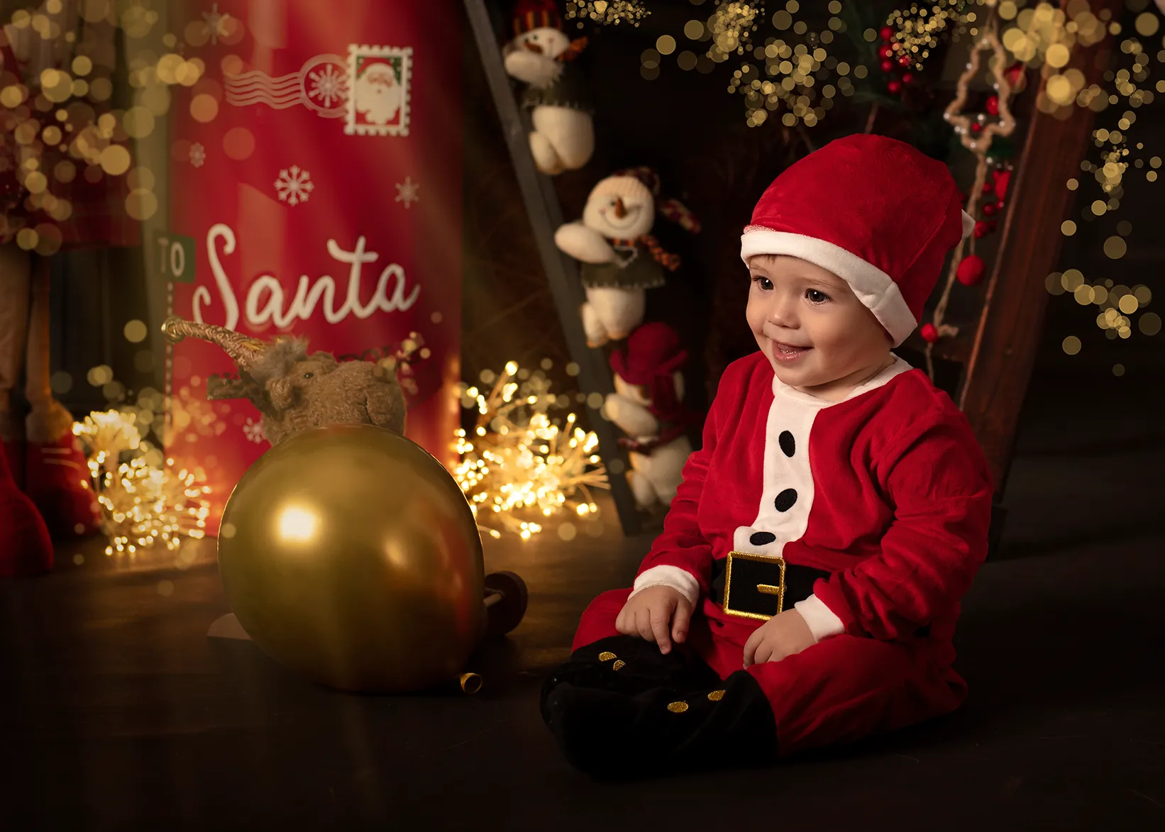 Un niño pequeño sonriente, vestido de Papá Noel, está sentado cerca de un globo dorado y adornos navideños.