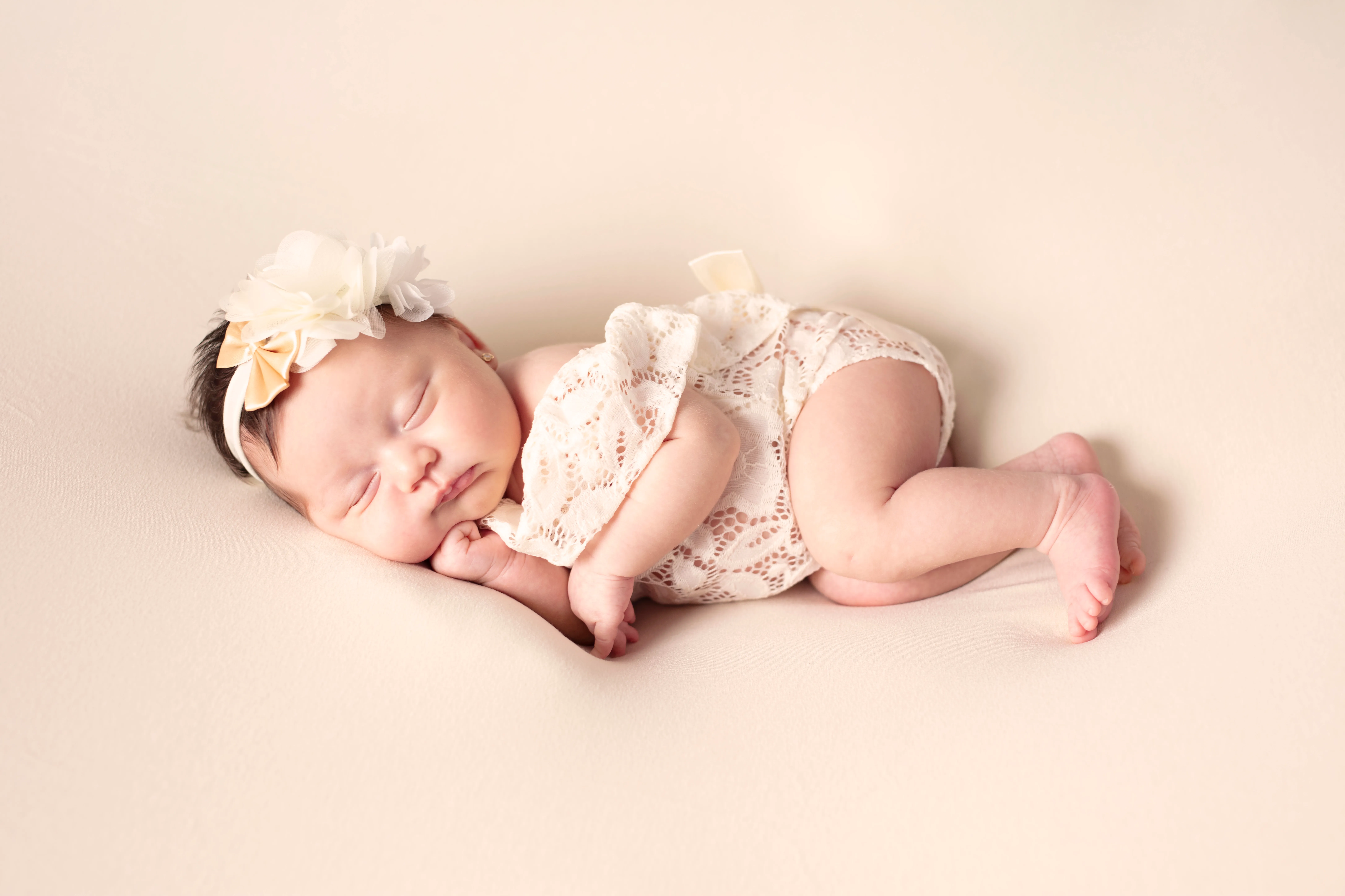 Bebé recién nacido durmiendo sobre una manta color crema, vestido con un conjunto de encaje y una diadema floral.