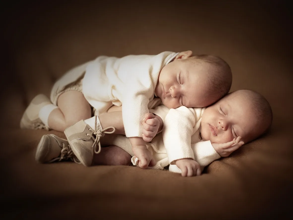 Dos bebés dormidos, vestidos con peleles blancos, sobre una manta marrón, uno encima del otro, con zapatitos.