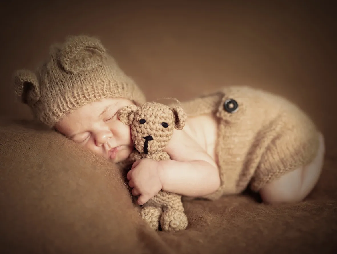 Bebé dormido con gorro y traje de oso marrón, sosteniendo un pequeño osito de peluche.