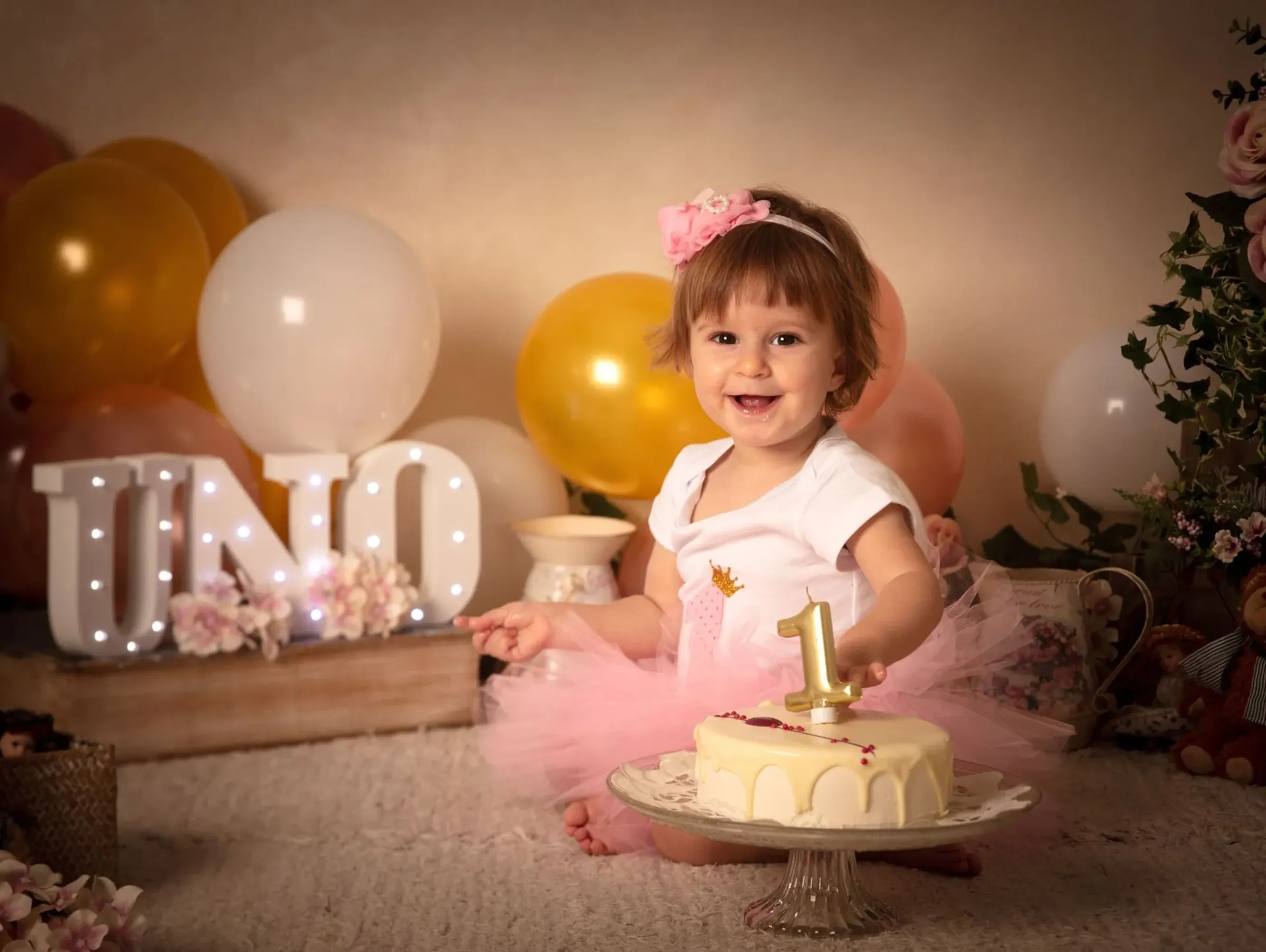 Niña sonriente celebrando su primer cumpleaños con pastel y el cartel de 