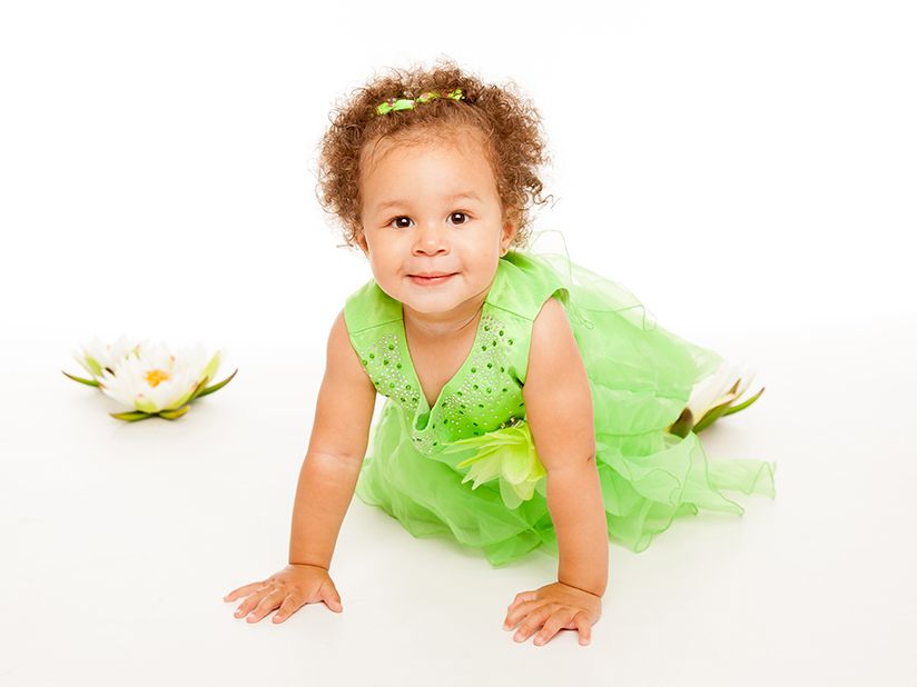 Una niña pequeña con un vestido verde gatea hacia la cámara, sonriendo, cerca de flores blancas sobre un fondo blanco.