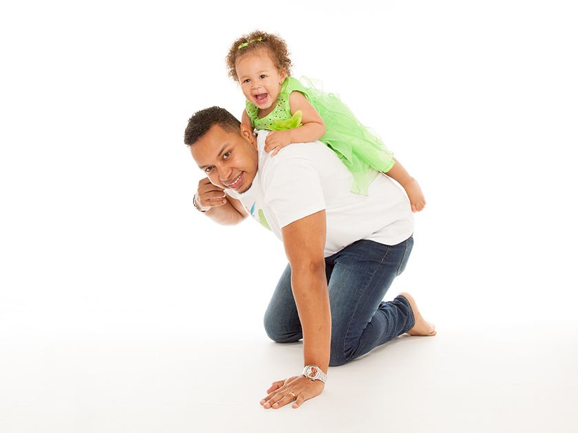 Hombre arrodillado con un niño a cuestas, ambos sonriendo frente a un fondo blanco.