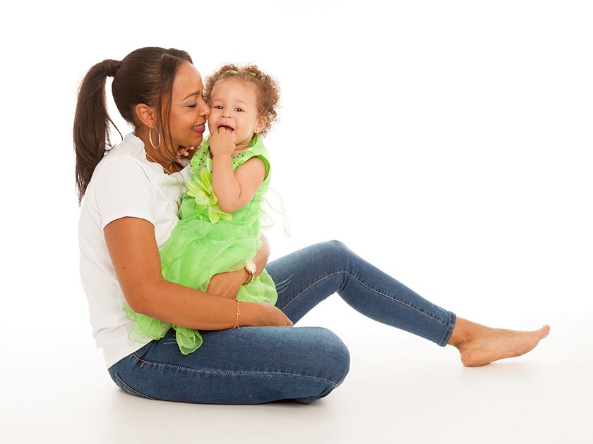Una mujer con camisa blanca y vaqueros está sentada en el suelo sosteniendo a una niña que ríe vestida con un vestido verde.