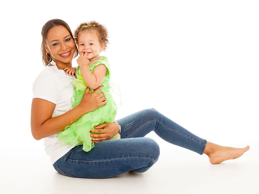 Mujer en vaqueros y camisa blanca sosteniendo a una niña con vestido verde, ambas sonriendo, sobre fondo blanco.