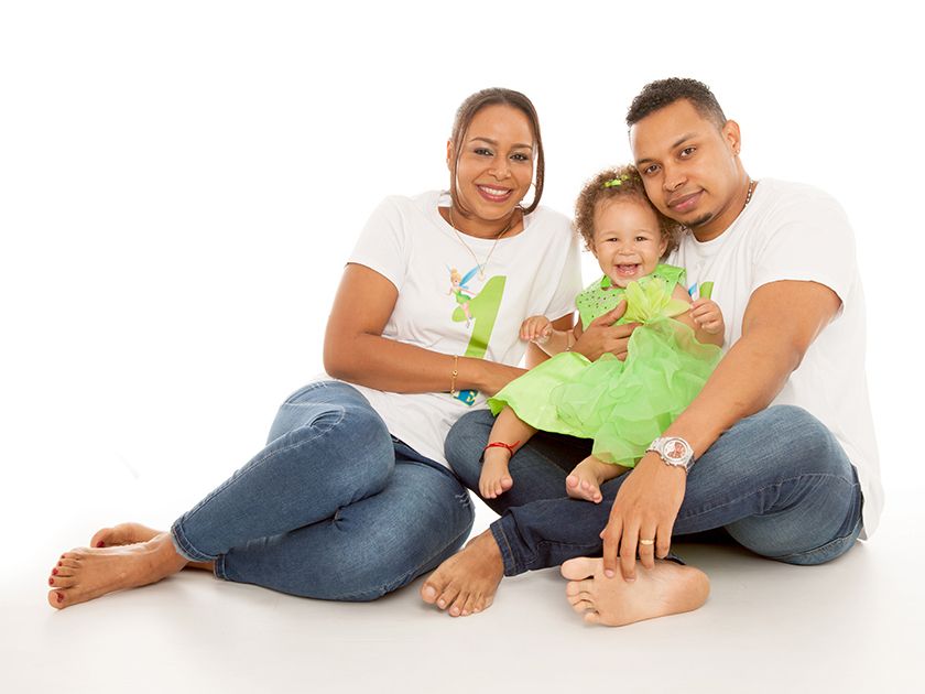 Familia de tres personas sentada, sonriendo, sobre un fondo blanco. Niña con vestido verde, padres con camisas blancas y vaqueros.