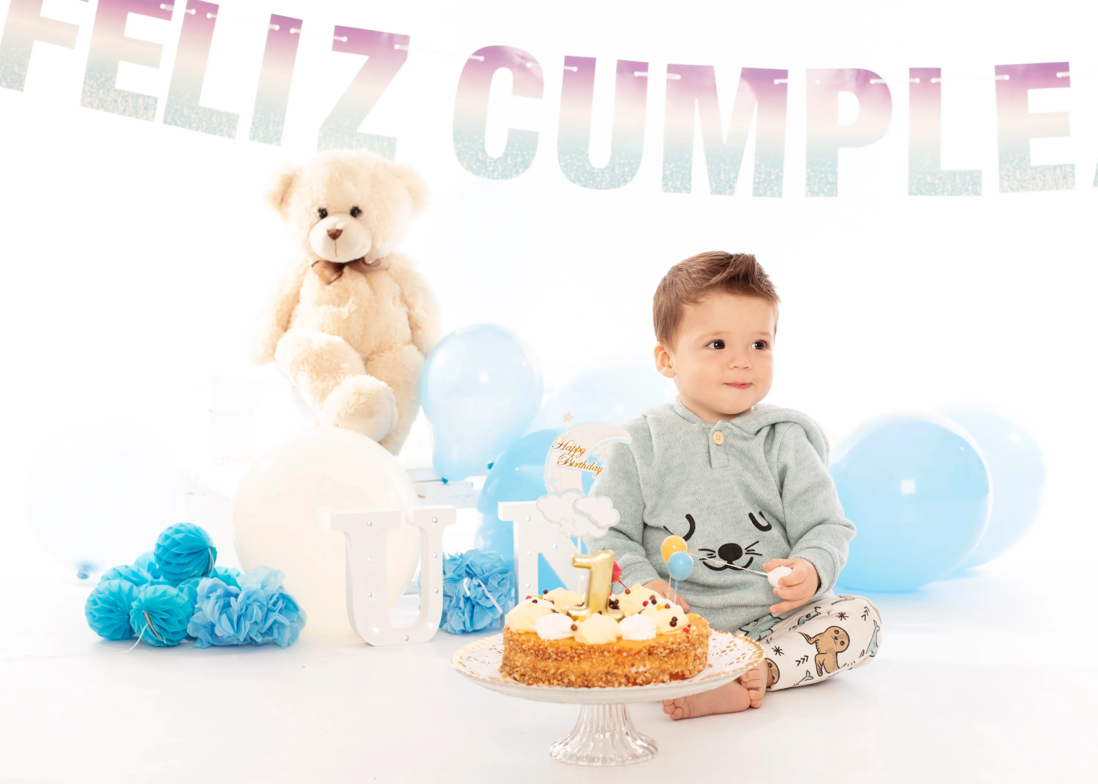 Niño sentado frente a un pastel de cumpleaños, rodeado de globos, un osito de peluche y una pancarta que dice 