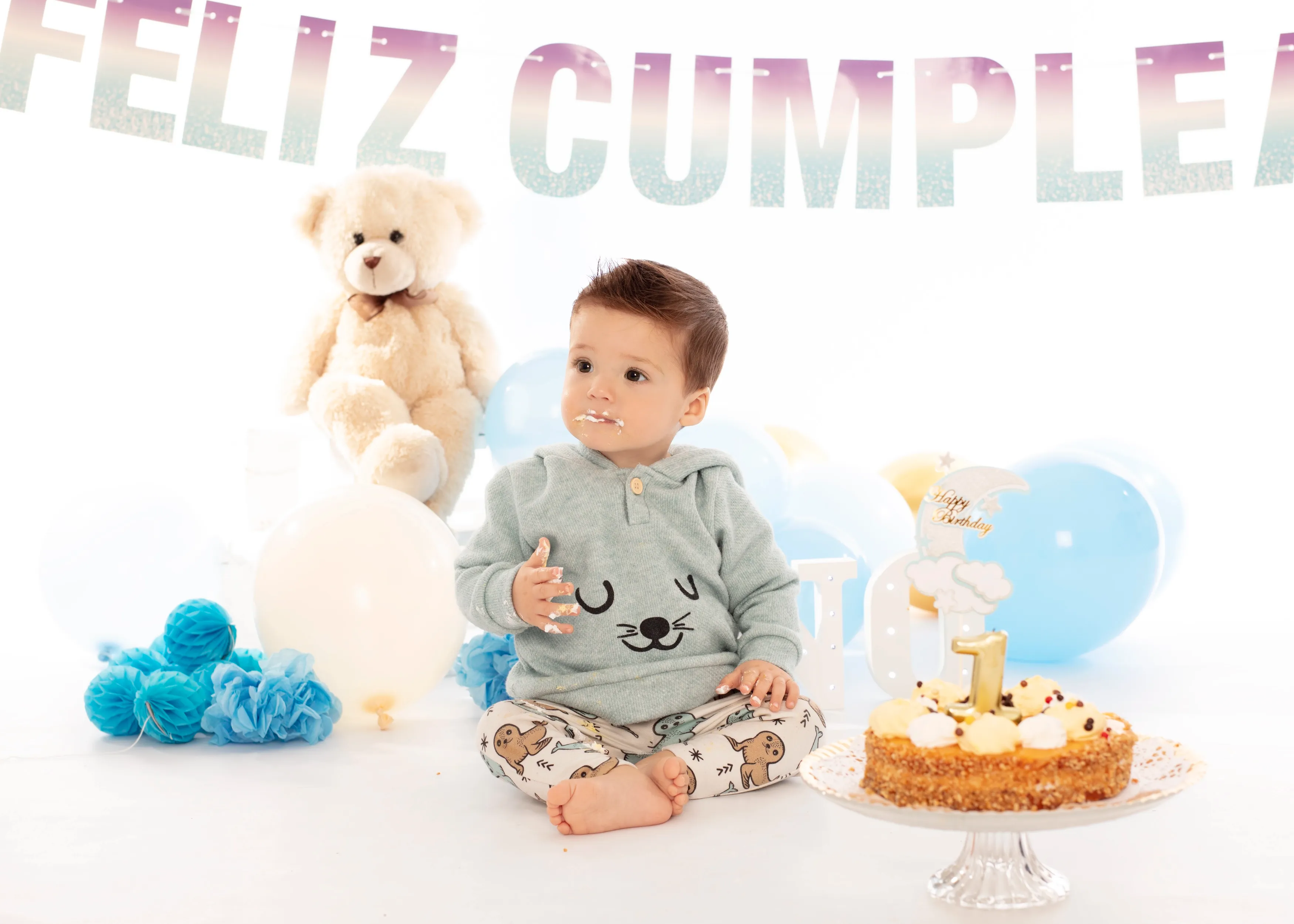 El bebé está sentado con pastel, globos, osito de peluche y una pancarta de “Feliz Cumpleaños”, celebrando su primer cumpleaños.