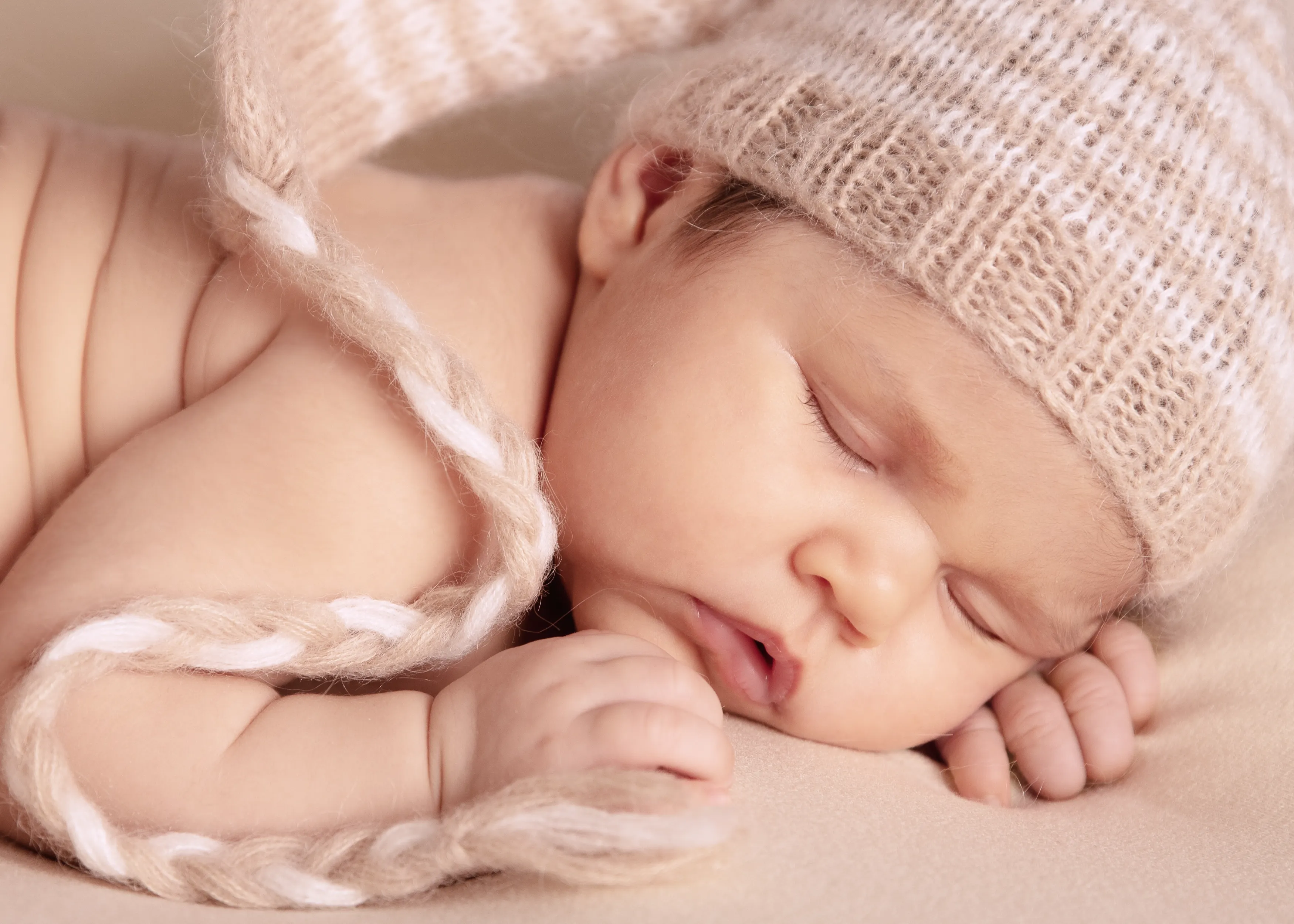 Bebé dormido con gorro de lana, descansando sobre una superficie beige con la cabeza apoyada en las manos.