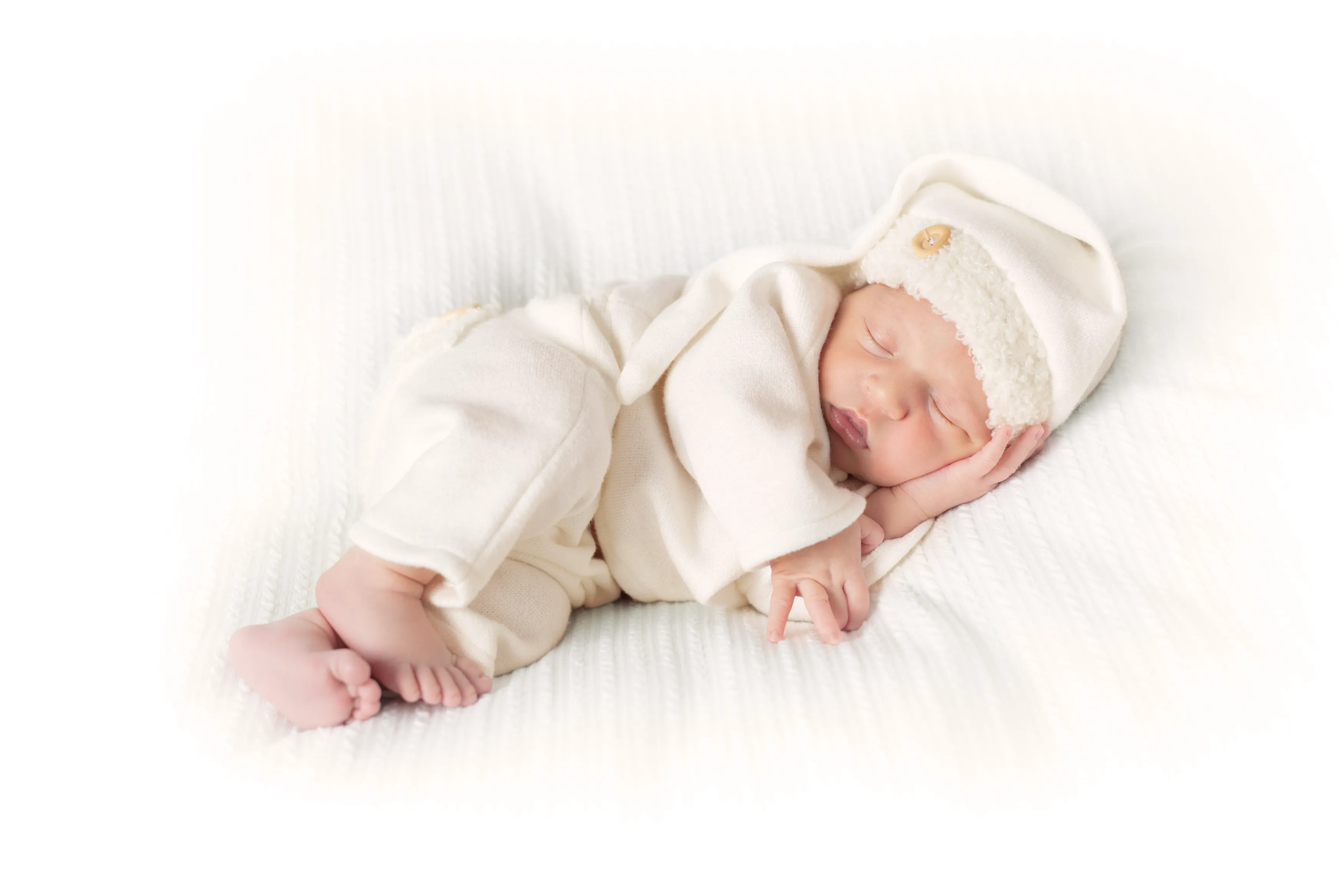 Bebé durmiendo con gorro y ropa blancos, tumbado sobre una manta blanca.