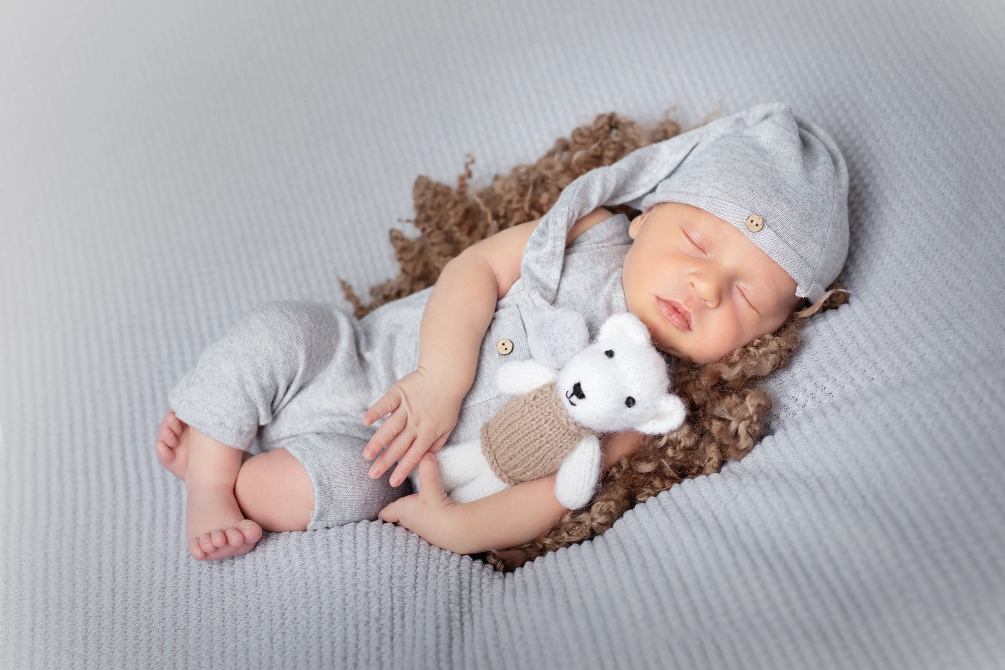 Bebé durmiendo con un conjunto de punto gris, acostado sobre una manta gris y sosteniendo un osito de peluche blanco.