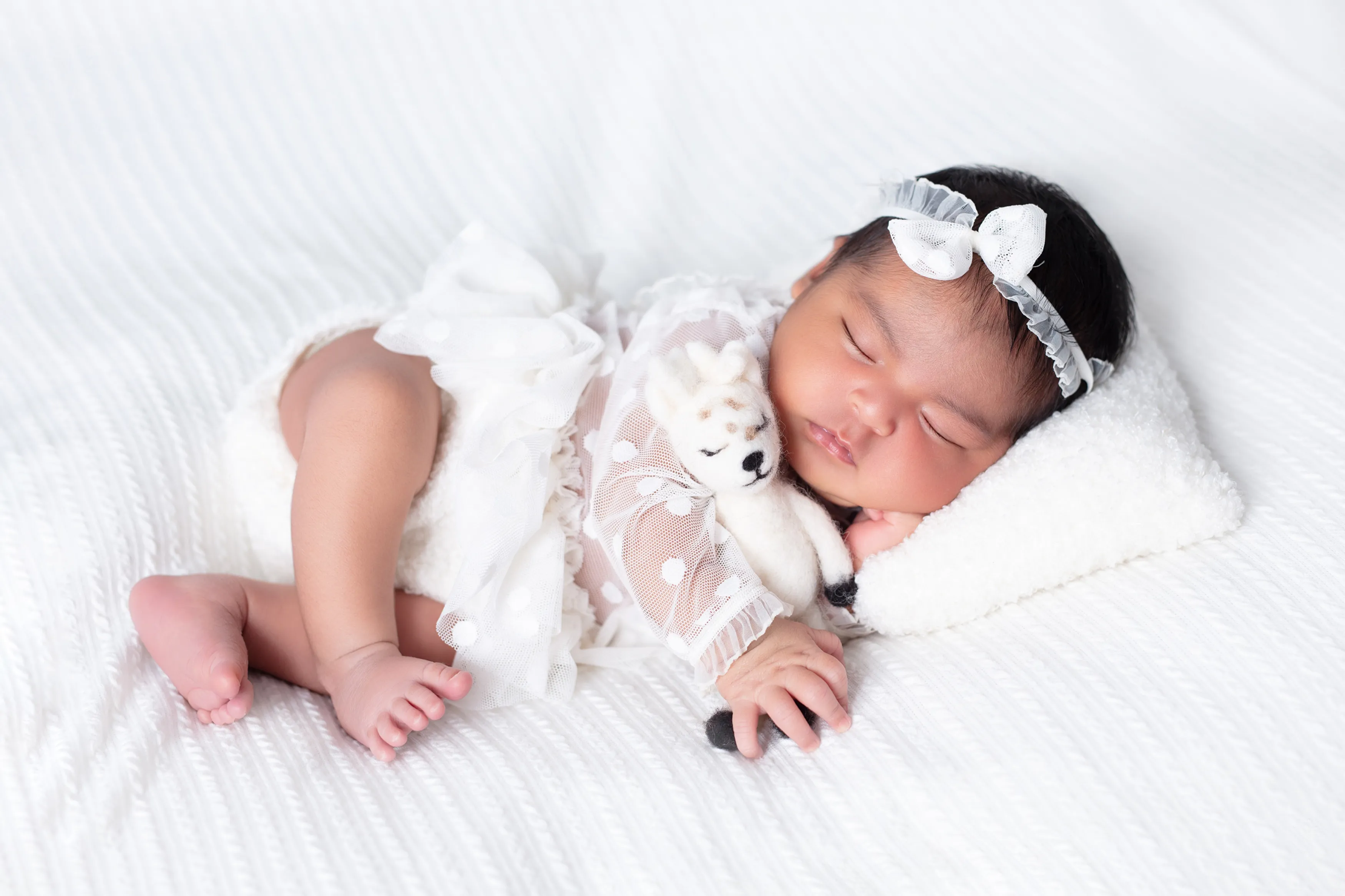 Bebé durmiendo con un conjunto de encaje blanco sobre una manta blanca, descansando sobre una pequeña almohada.