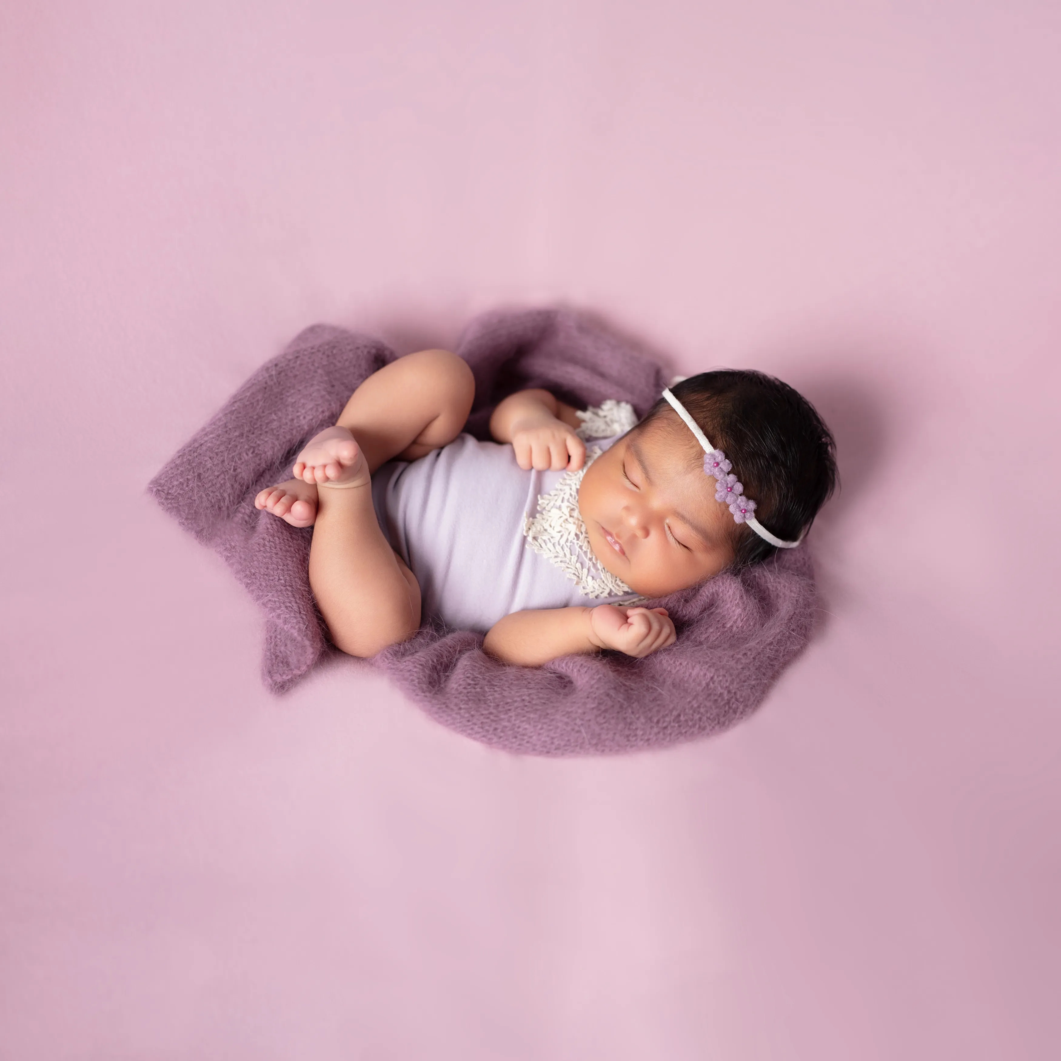 Recién nacido durmiendo envuelto en una manta color lavanda sobre un fondo rosa.