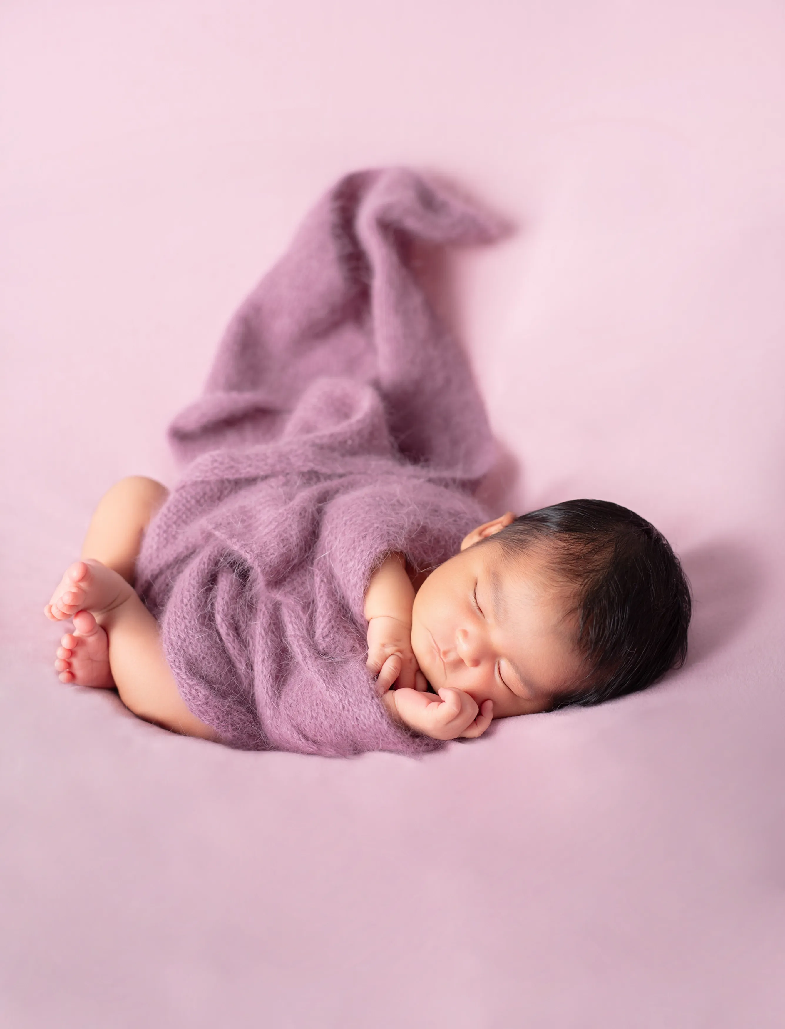 Bebé durmiendo envuelto en una manta color lavanda sobre un fondo rosa suave.