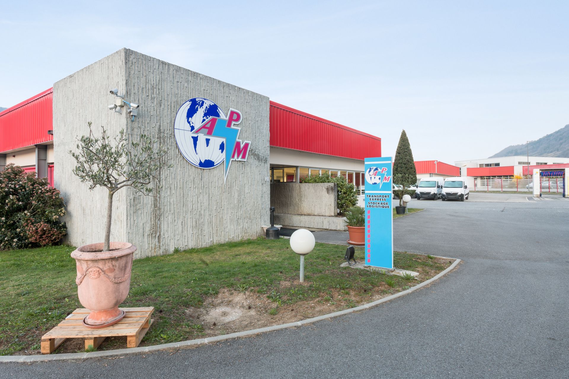 Bâtiment moderne avec toit rouge et cour intérieure, enseigne bleue, plante en pot et voitures garées sous un ciel dégagé