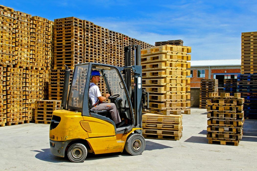 Un chariot élévateur jaune déplace des palettes dans une scierie, avec des piles de palettes en bois en arrière-plan.