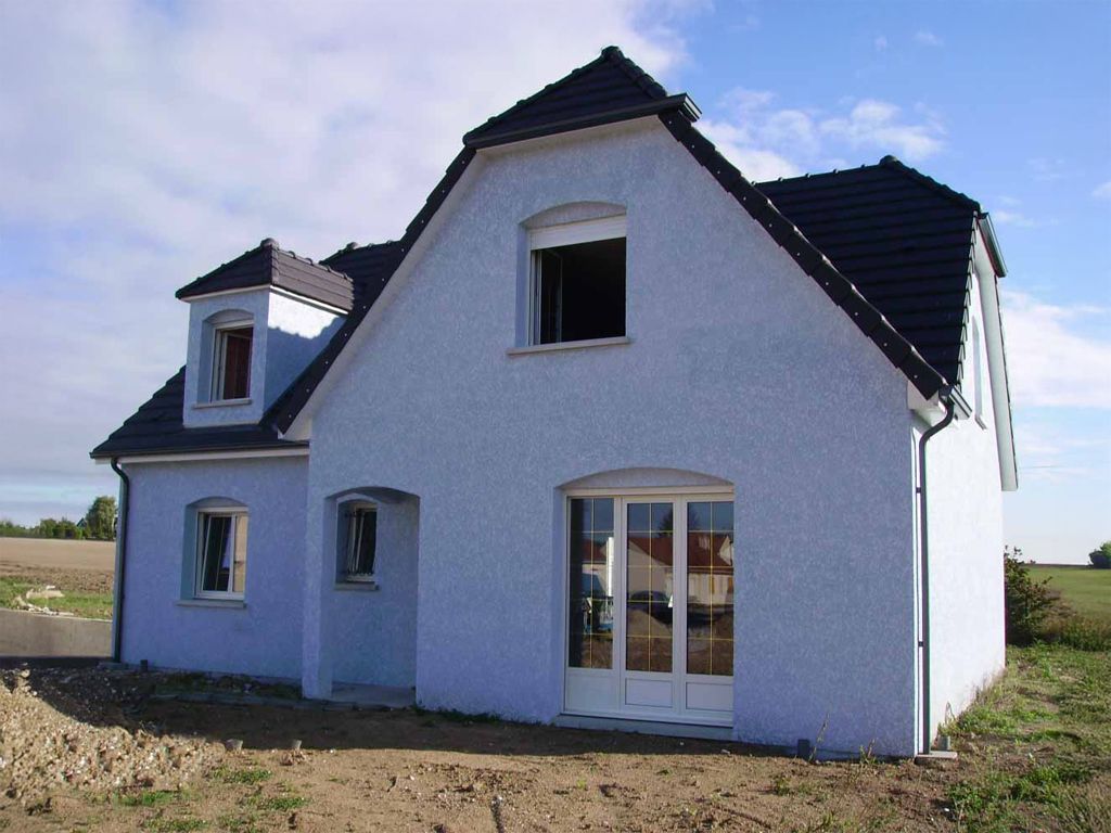 Maison bleu clair avec toit noir, fenêtres blanches et porte-fenêtre ; située dans un champ.