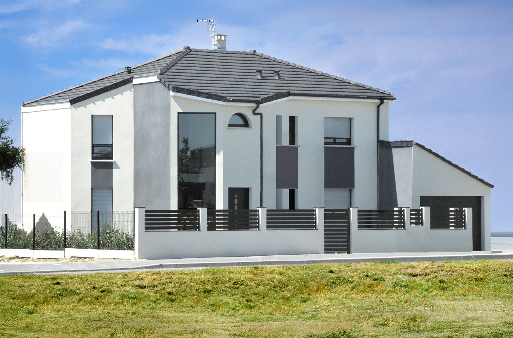 Maison blanche moderne à deux étages avec un toit et des accents gris, une cour clôturée et un petit garage.