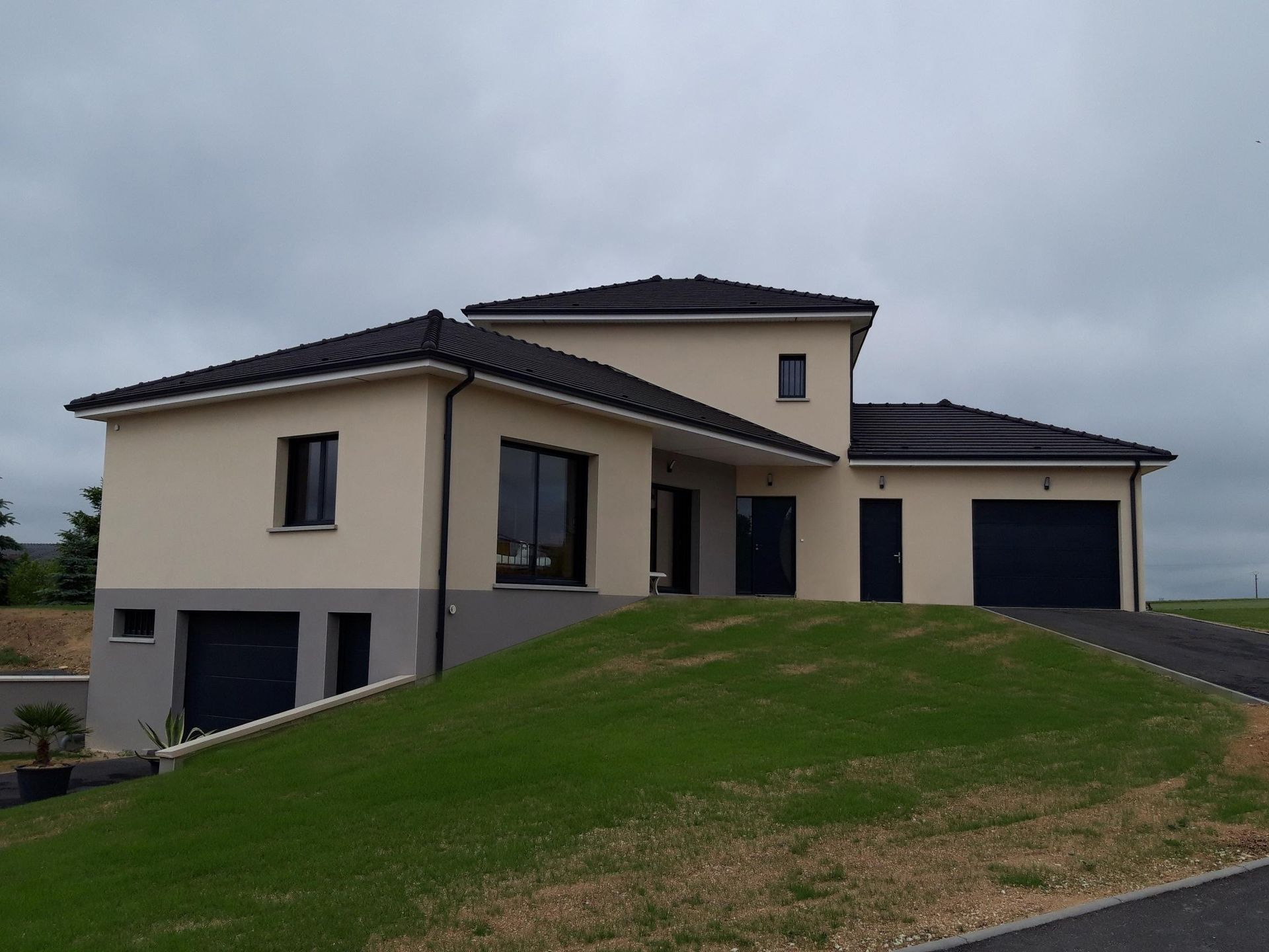 Maison beige moderne avec toit noir et portes de garage sur une colline herbeuse sous un ciel nuageux.