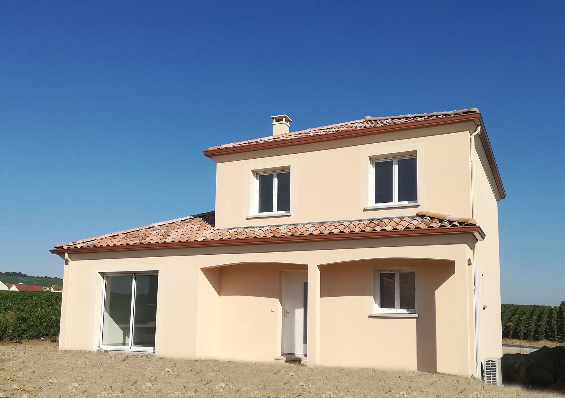 Maison beige à deux étages avec toit en tuiles rouges, cadres de fenêtres blancs, sous un ciel bleu clair.