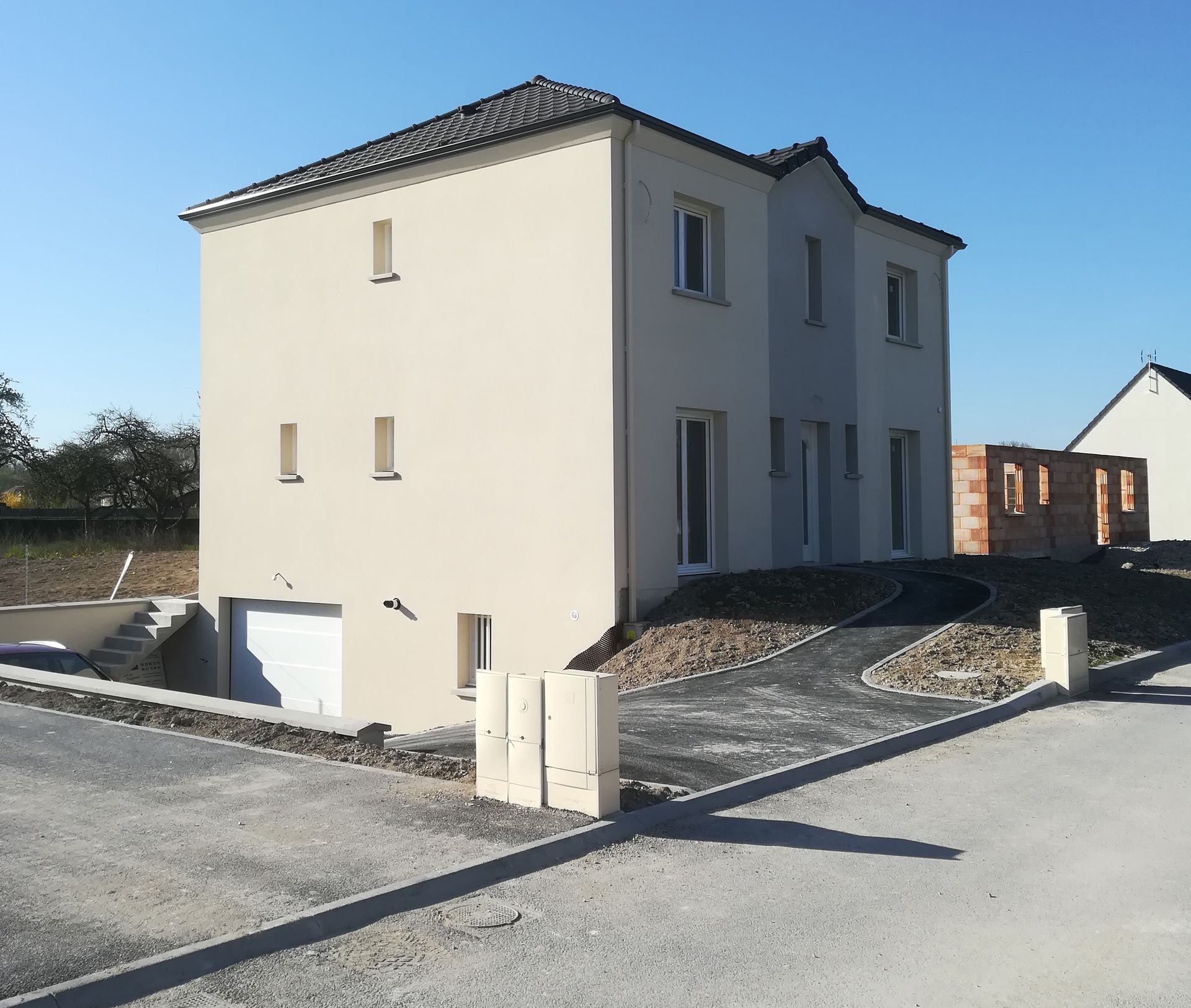 Maison beige à deux étages en construction, avec un garage, une allée et un autre bâtiment en arrière-plan.