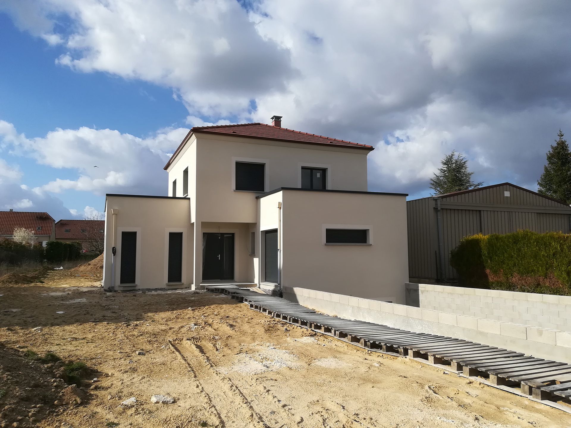 Maison beige moderne à deux étages sous un ciel bleu nuageux, avec aménagement paysager inachevé et un chemin.
