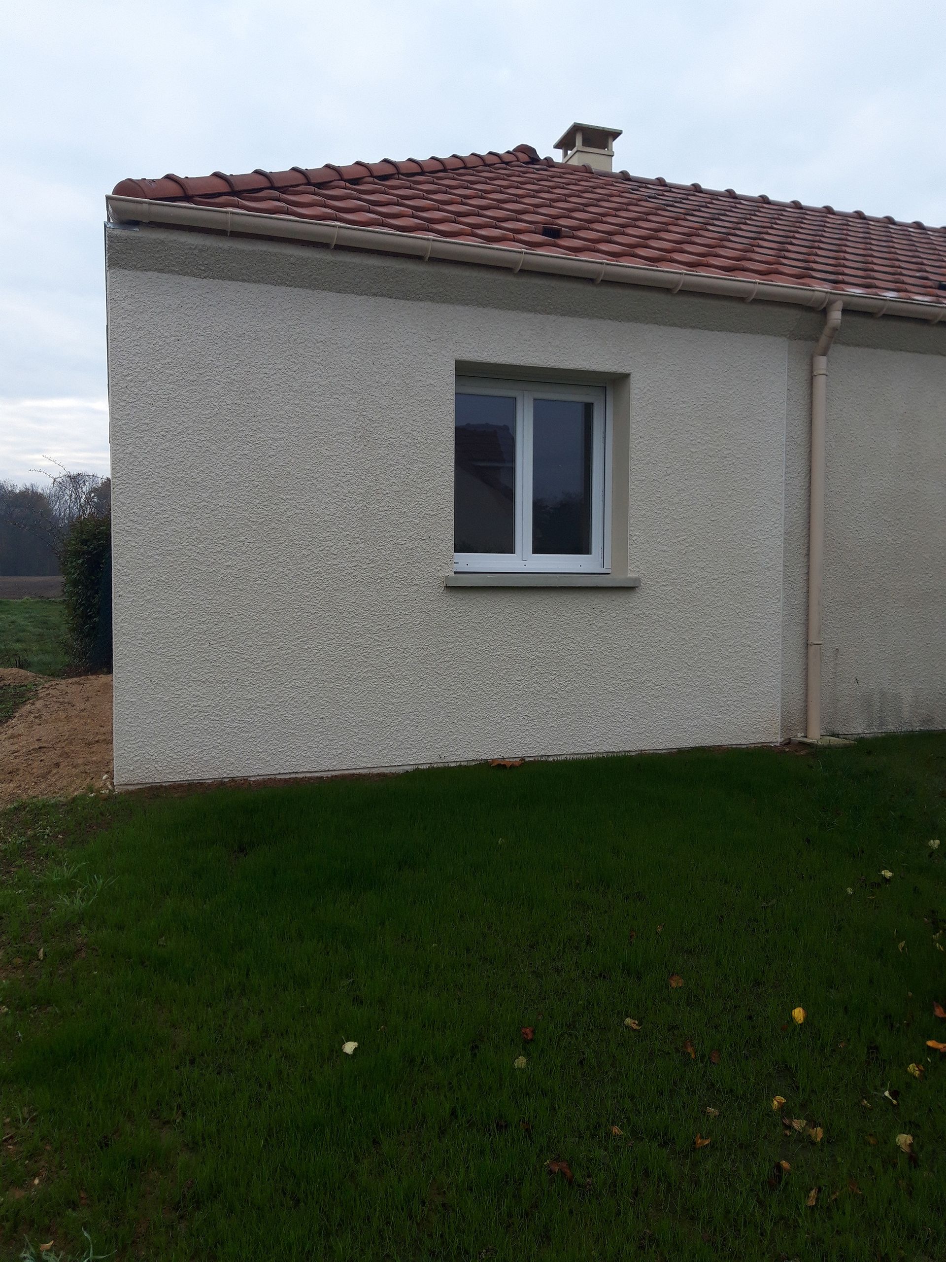 Une maison en stuc avec un toit de tuiles rouges, une fenêtre et de l'herbe verte devant. Ciel nuageux.
