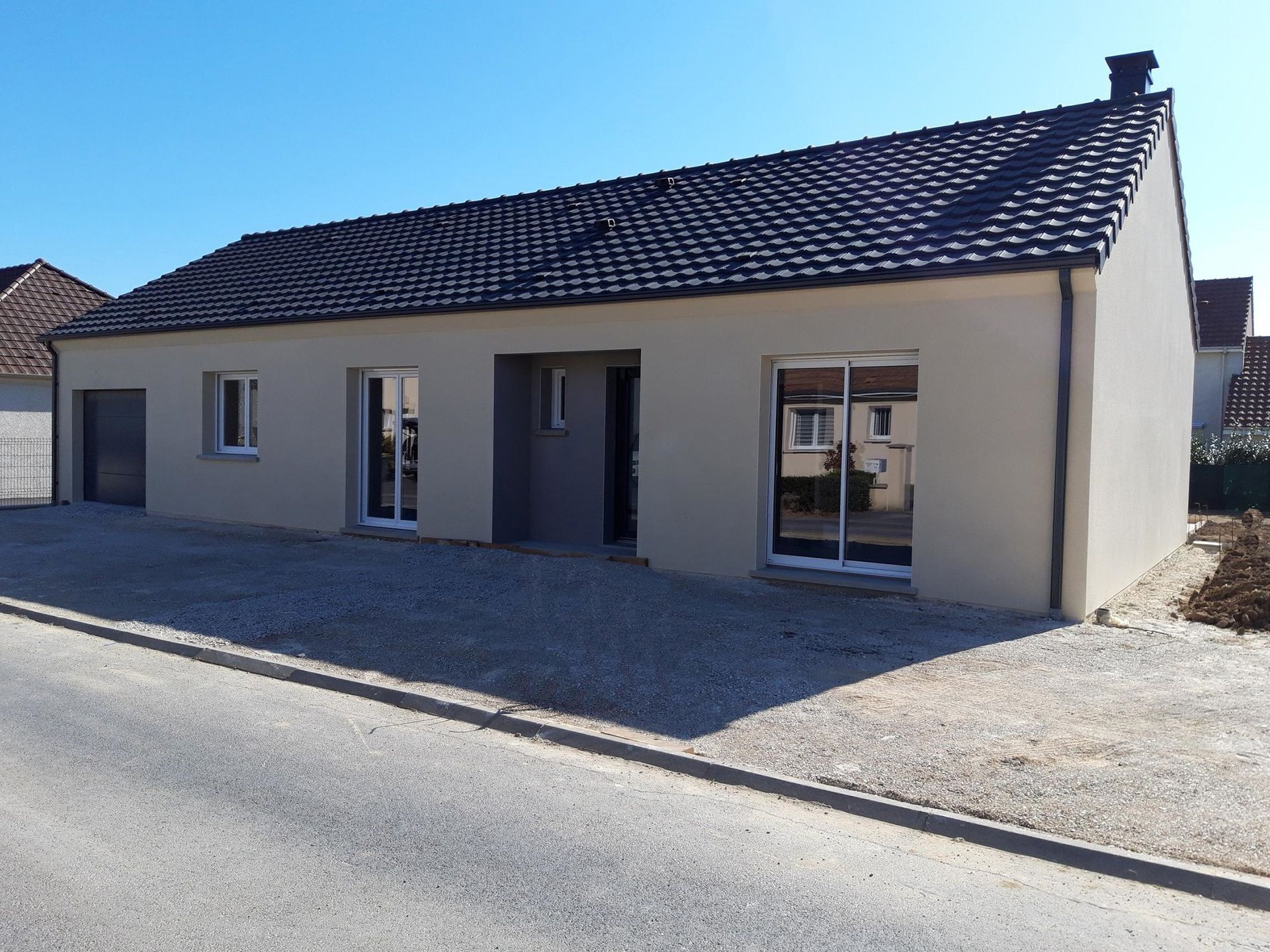 Maison beige avec toit noir, cadres de fenêtres blancs et allée de gravier gris, sous un ciel bleu.
