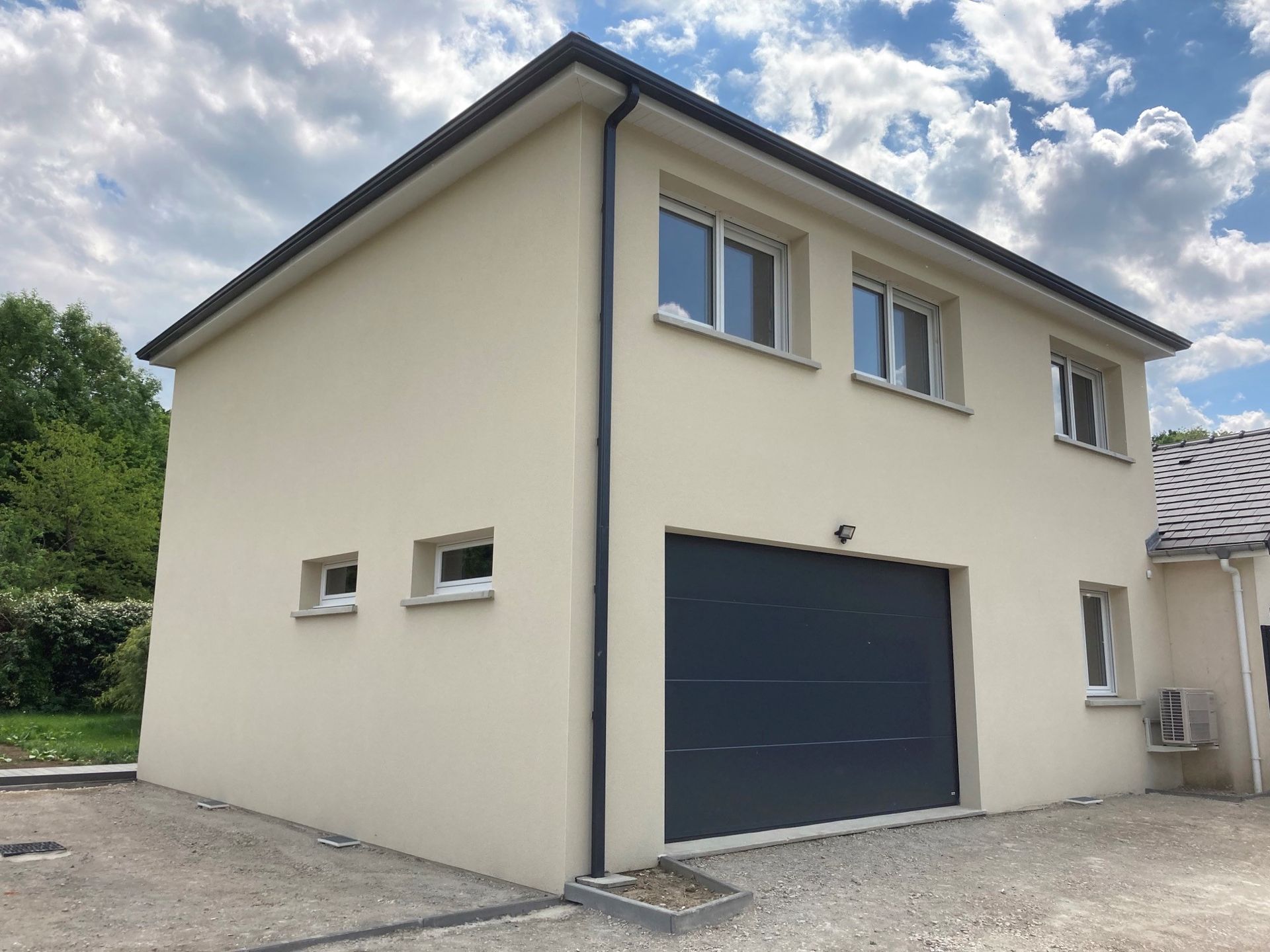 Maison beige à deux étages avec une porte de garage et des fenêtres gris foncé, située sous un ciel nuageux.