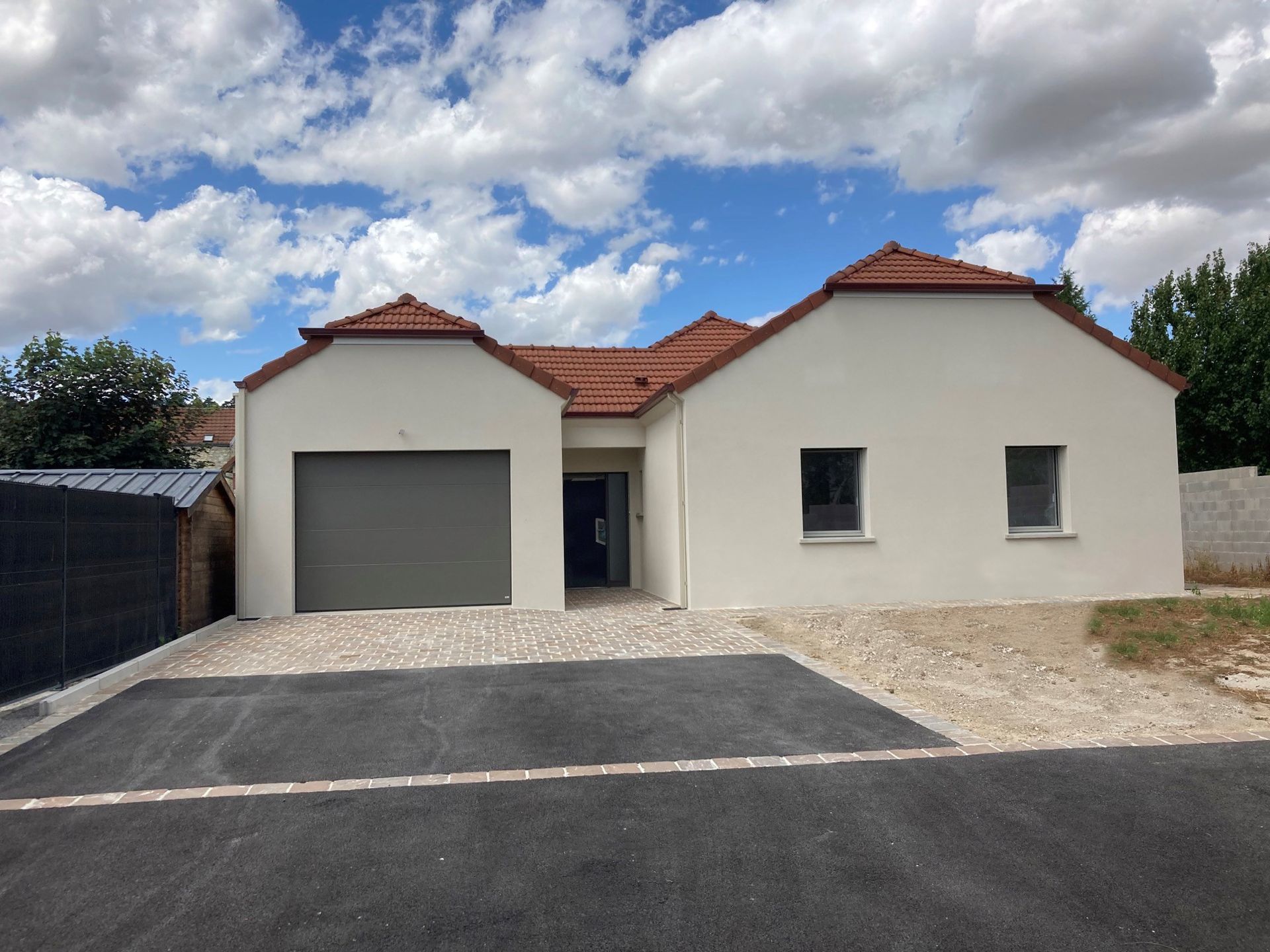 Vue extérieure d'une maison beige de plain-pied avec une porte de garage grise, un toit en tuiles rouges et un ciel bleu.
