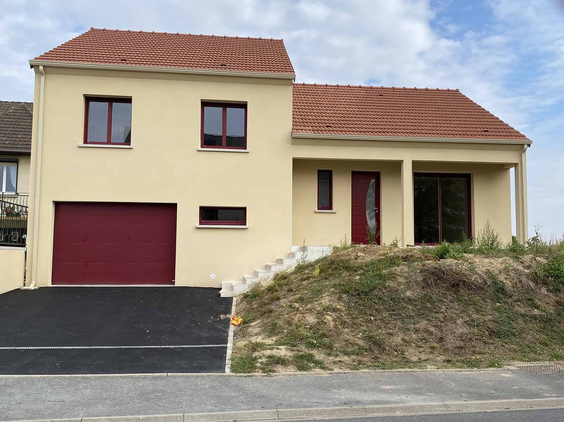 Maison beige à deux étages avec porte de garage et porte d'entrée marron, toit en tuiles rouges et pente herbeuse.