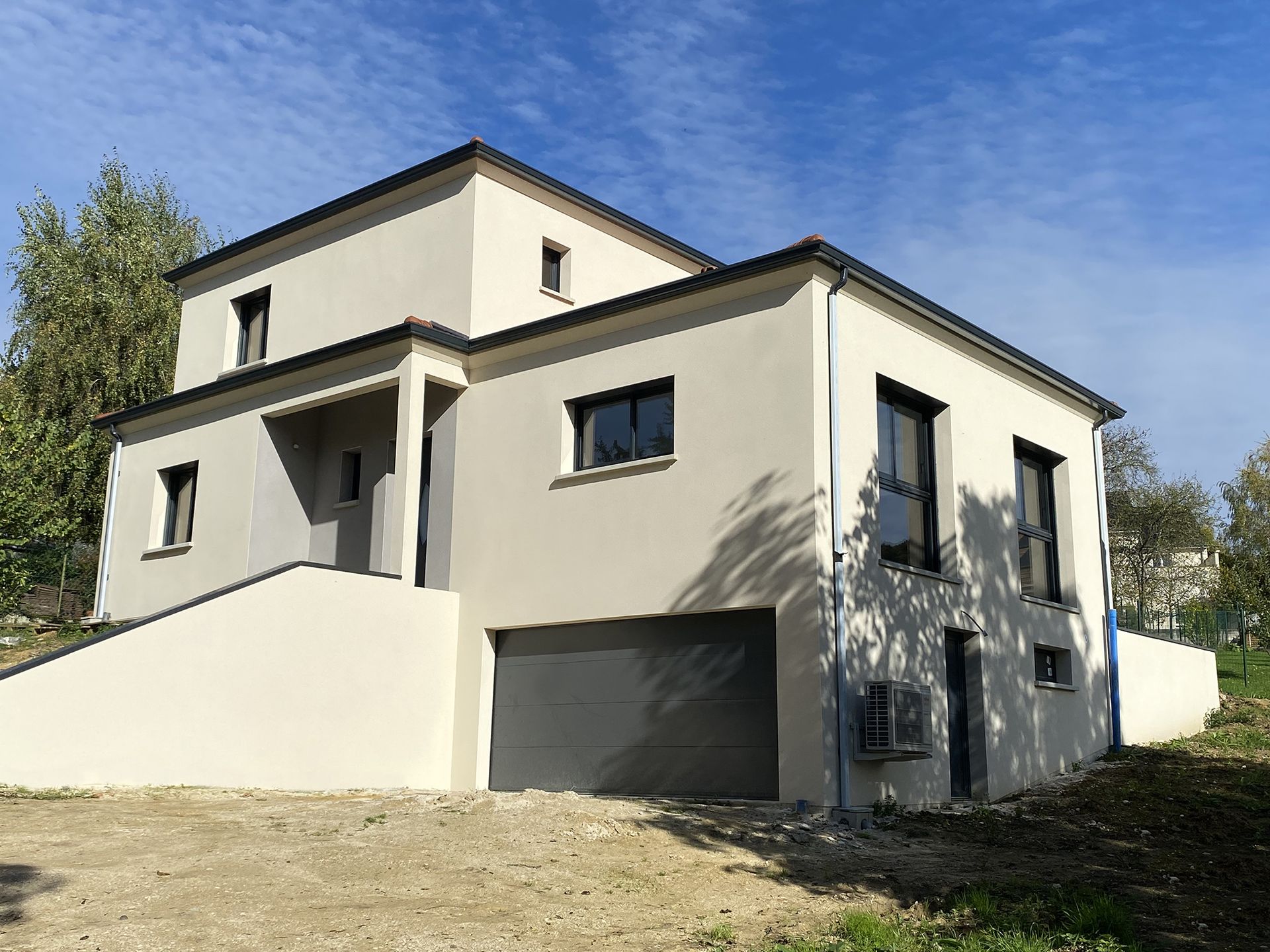 Maison moderne à deux étages avec stuc gris clair, toit foncé et garage. Nichée sur une légère colline sous un ciel bleu.