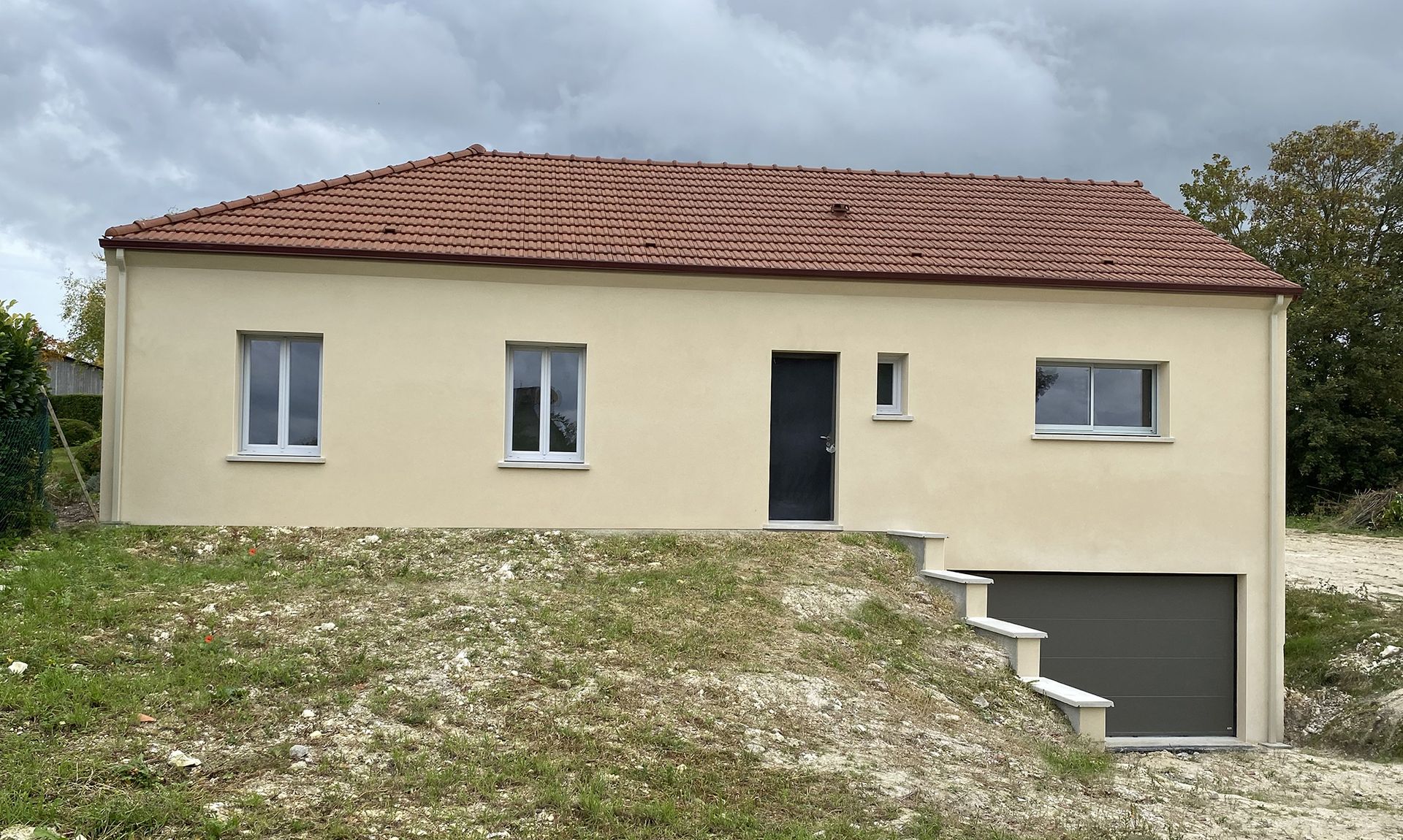 Maison beige avec toit en tuiles rouges, porte d'entrée, garage et pelouse.