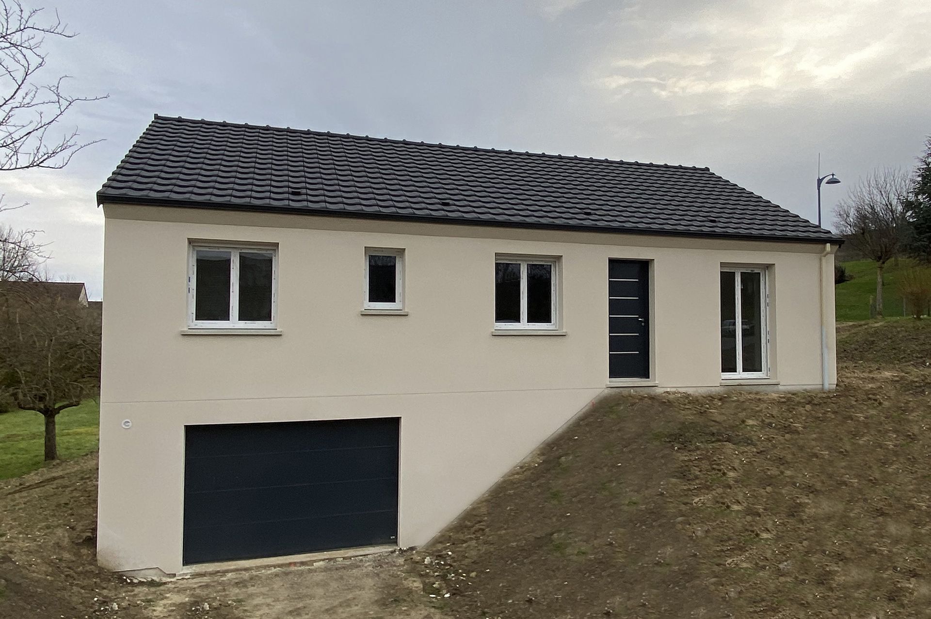 Maison beige au toit noir et garage. Située sur une petite colline, elle possède des fenêtres et une porte sombres.