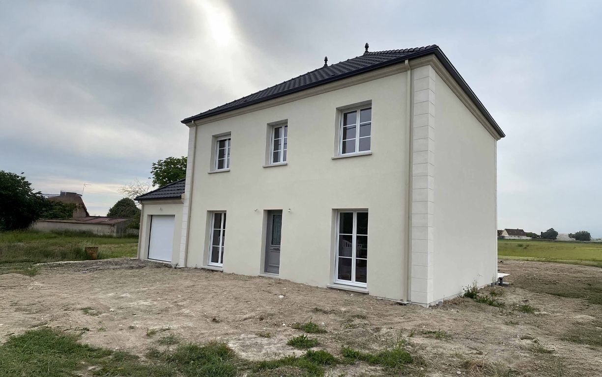 Maison beige à deux étages avec fenêtres à cadres blancs, toit noir et garage. Située sur un terrain poussiéreux.