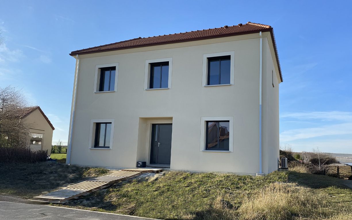 Maison beige à deux étages avec fenêtres et porte sombres, toit en tuiles rouges, assise sur une petite colline herbeuse.