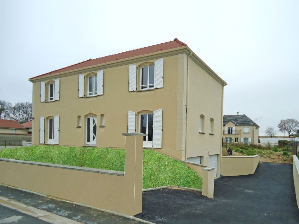 Maison beige à deux étages avec volets blancs et toit rouge ; talus gazonné devant, ciel couvert.