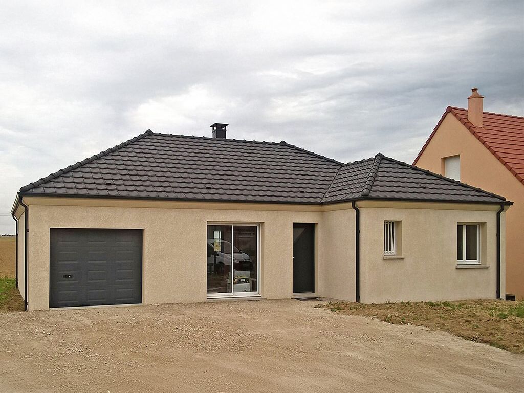 Maison beige de plain-pied avec toit foncé, garage et allée en gravier, par temps couvert.