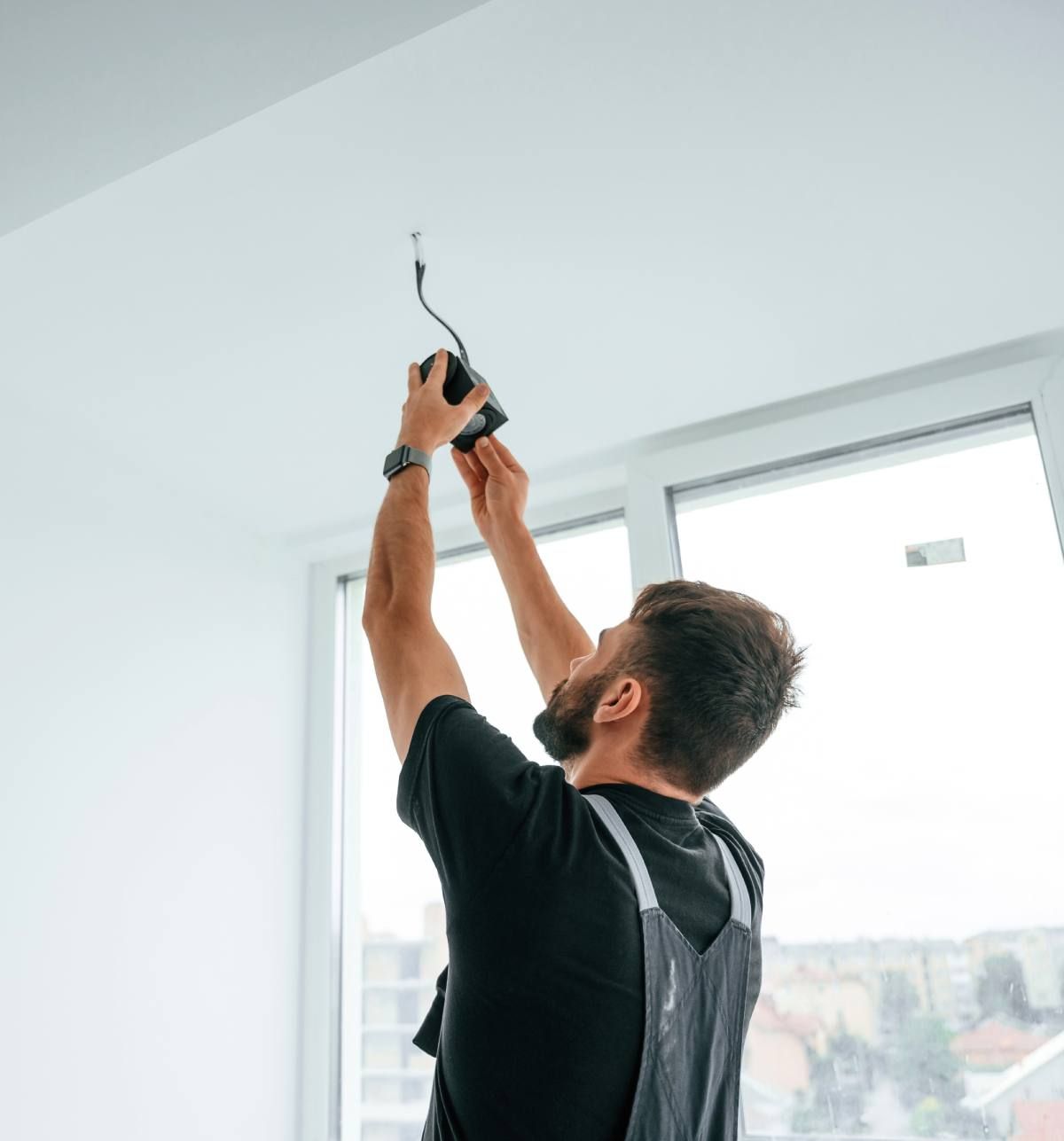Un homme en salopette installe un luminaire sur un plafond blanc près d'une fenêtre.