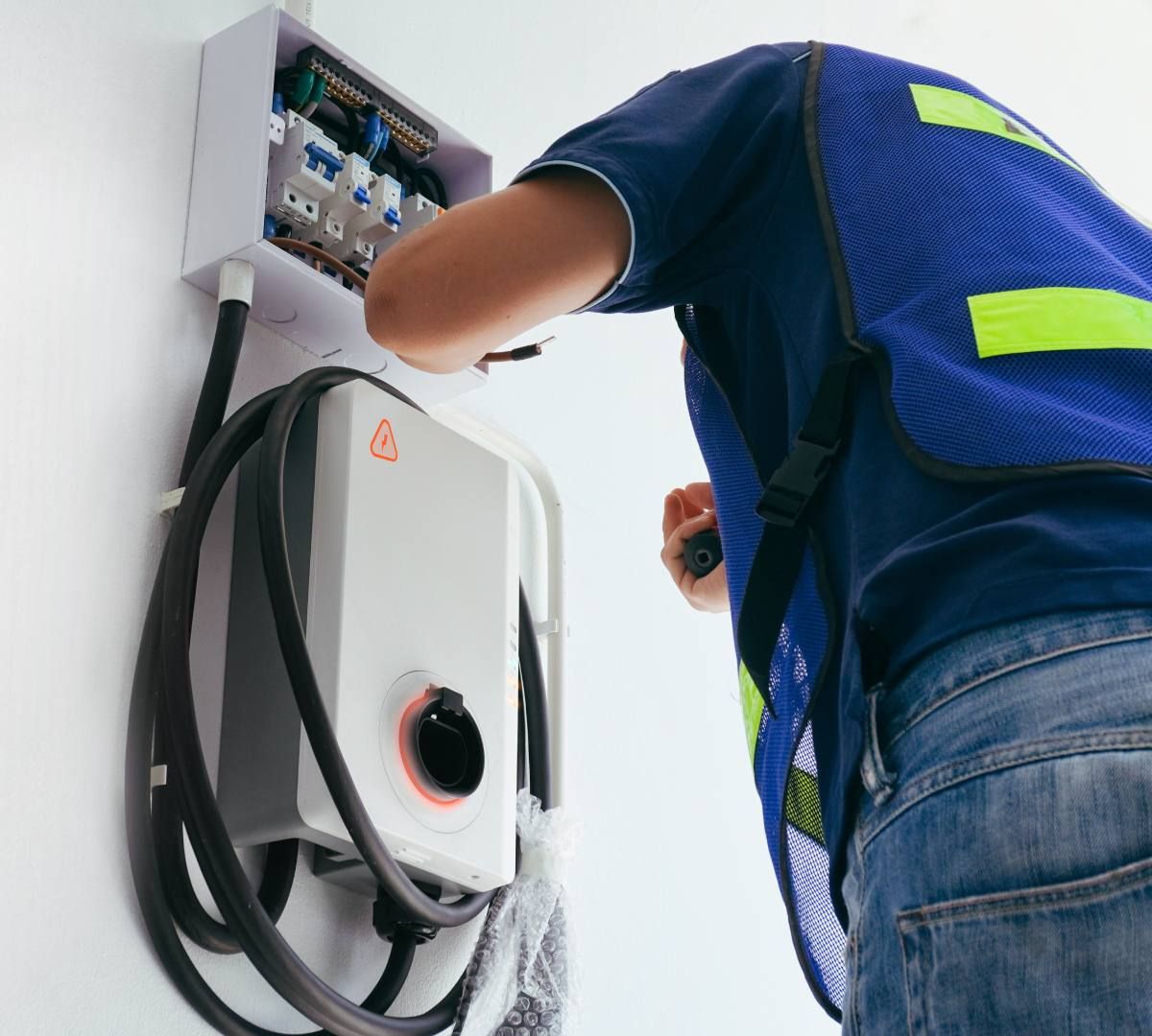 Électricien installant un chargeur de VE sur un mur blanc ; gilet bleu, panneau électrique visible.