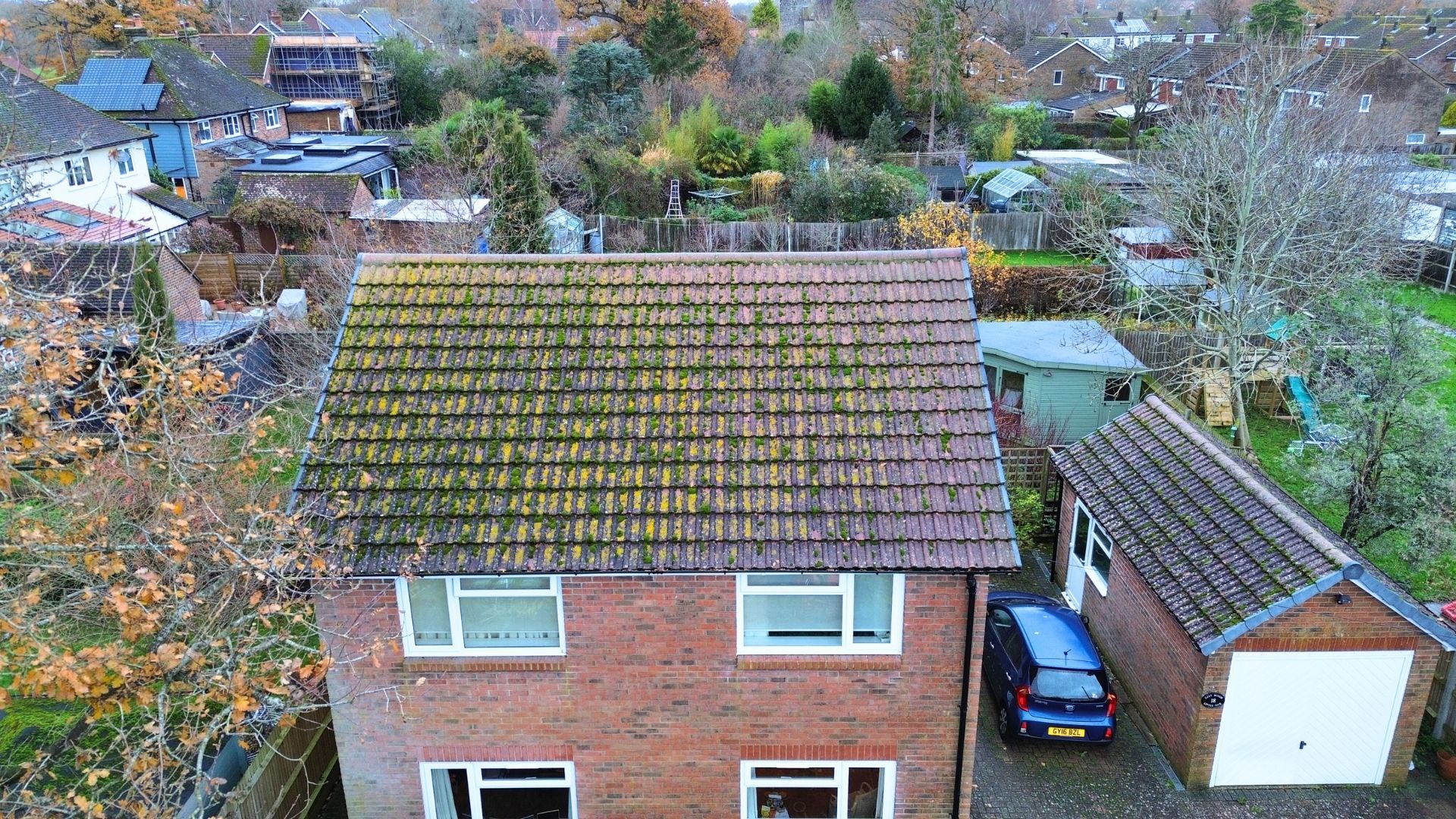 Aerial view of a brick house with a mossy roof, driveway, parked car, and a white shed in a residential area